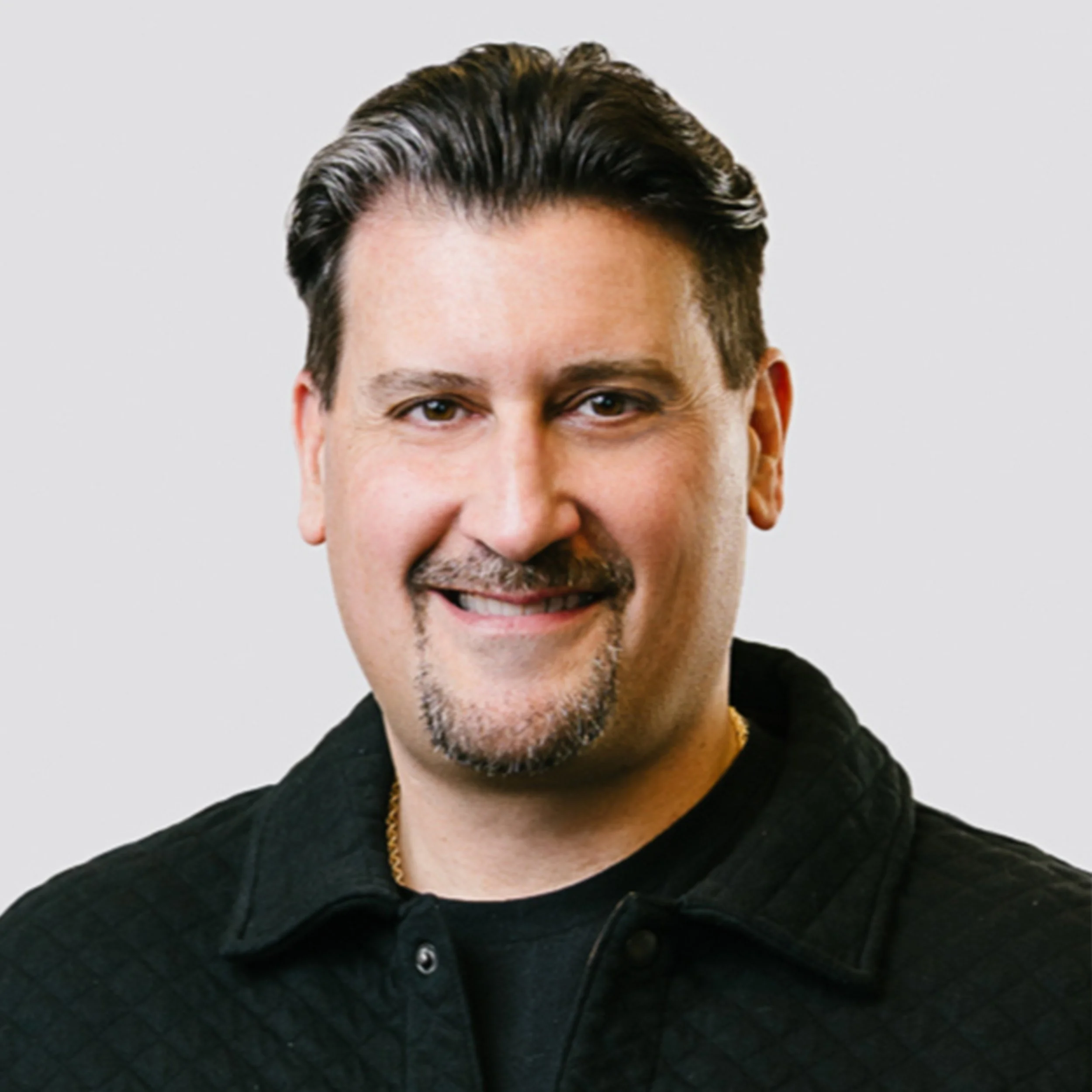 Headshot of a smiling man with dark hair, a beard, and a goatee, wearing a black quilted shirt against a plain light-gray background.
