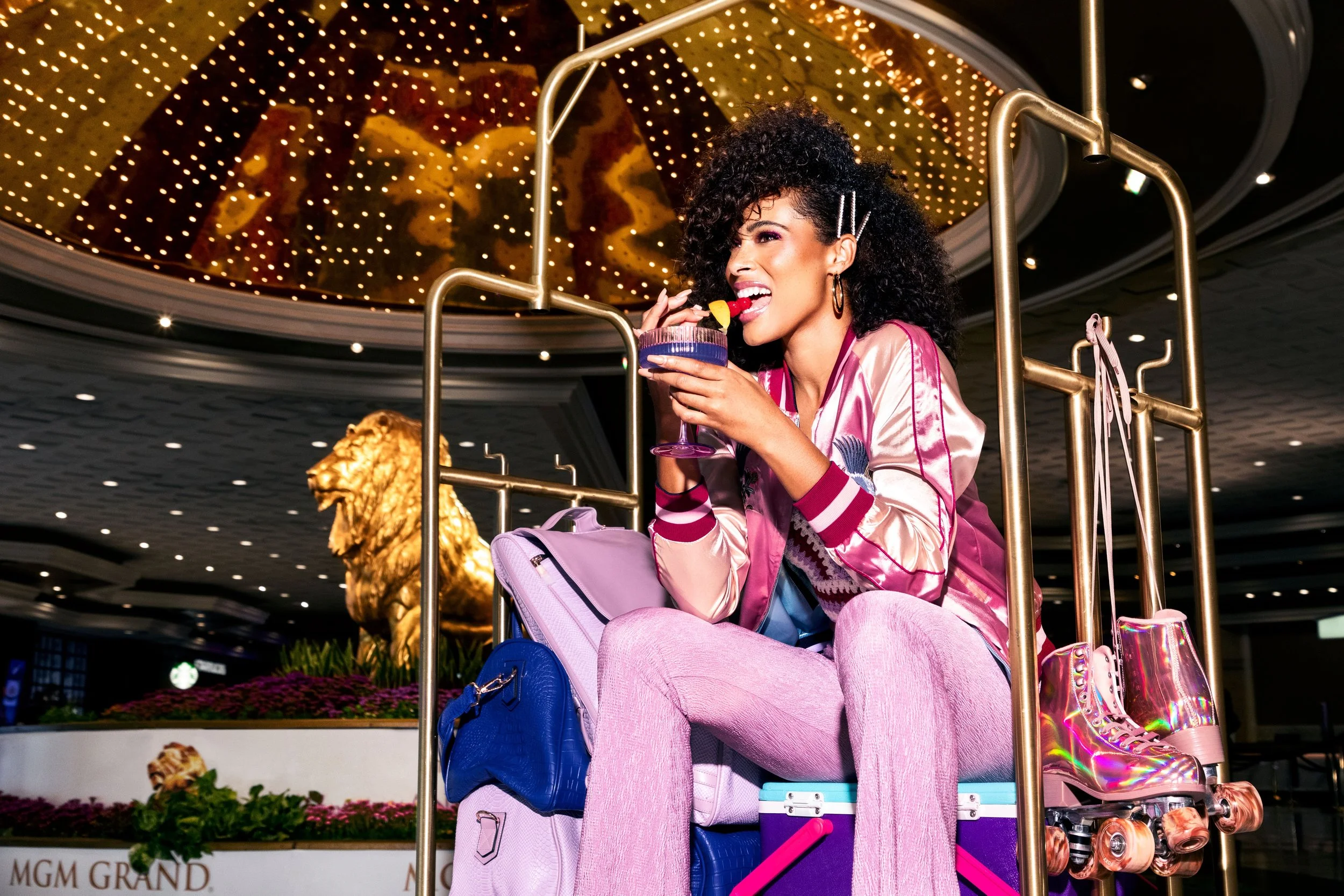 A woman with curly hair sitting on a luggage cart inside a casino or hotel, licking a colorful cocktail, with roller skates attached to the cart, and a large gold lion statue in the background.