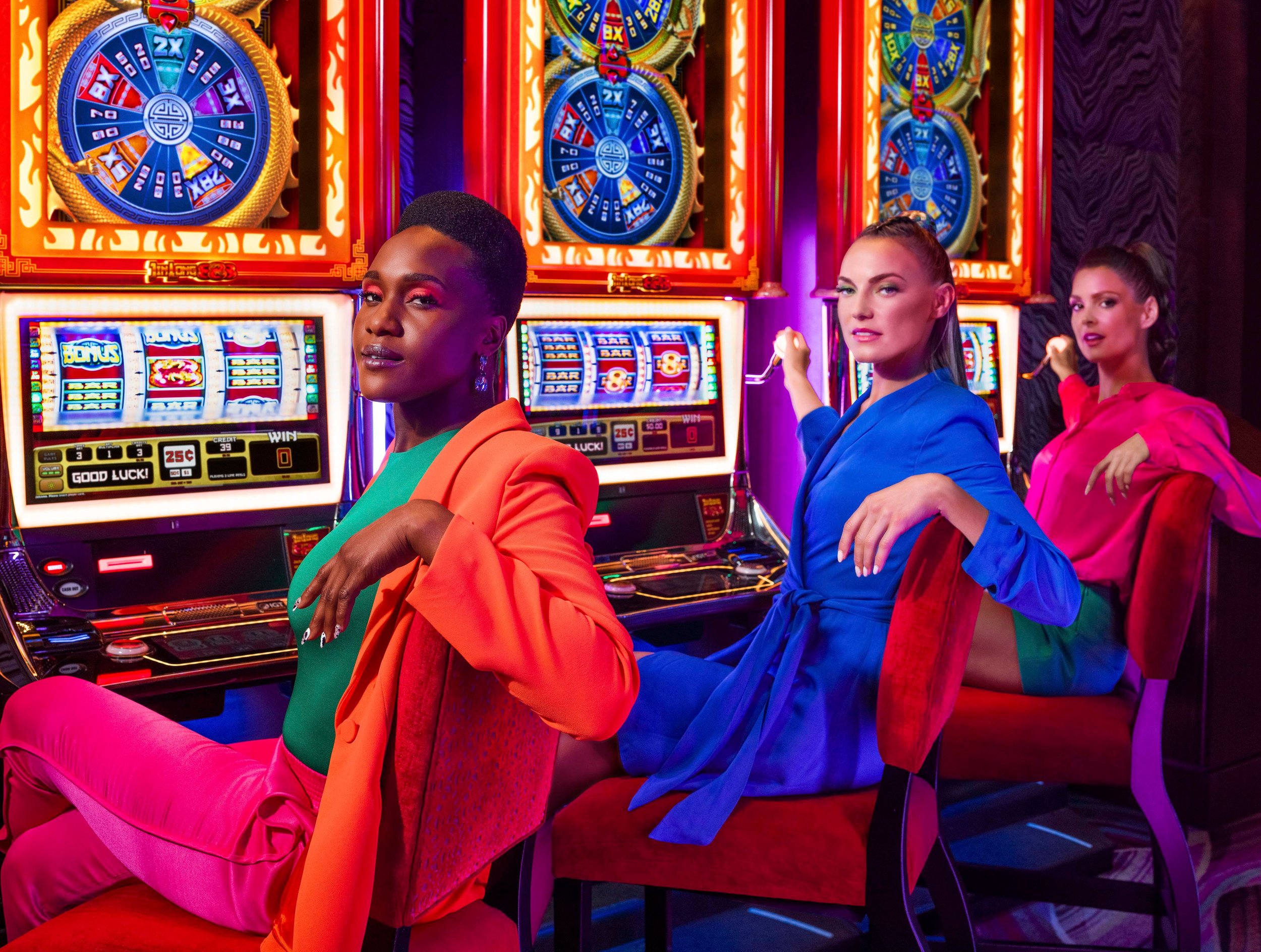 Three women sitting in front of slot machines in a brightly lit casino, dressed in colorful outfits, with colorful lights and gaming displays in the background.