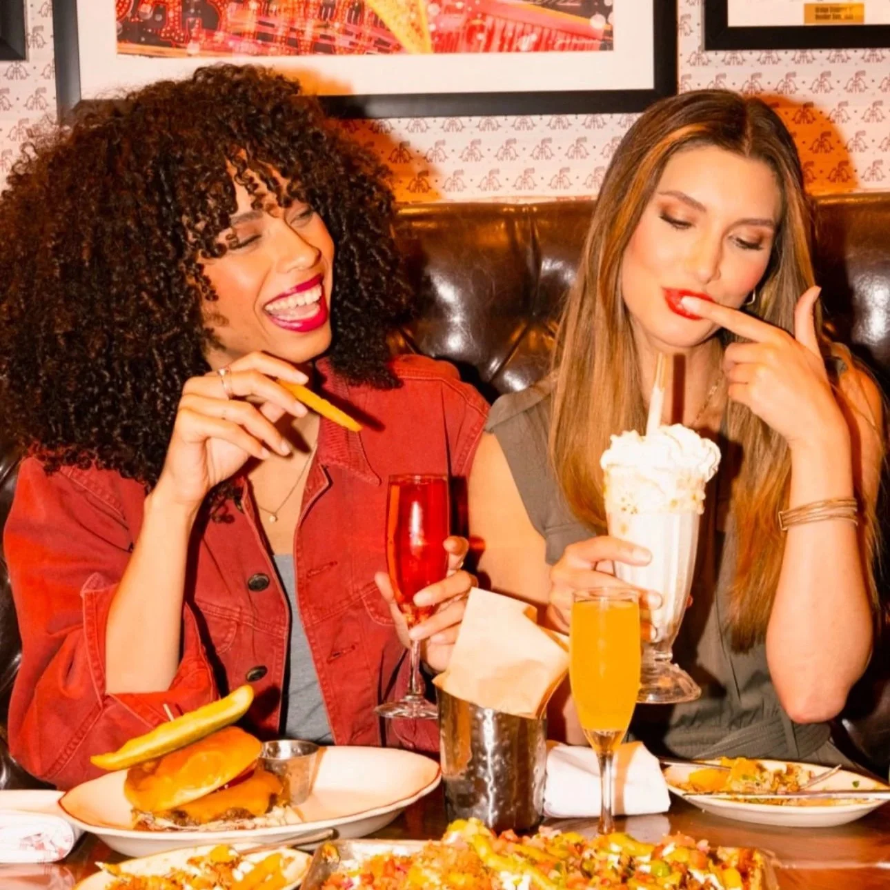 Two women sitting at a restaurant table enjoying food and drinks, one with curly hair and the other with straight hair, with plates of pizza and salad on the table.