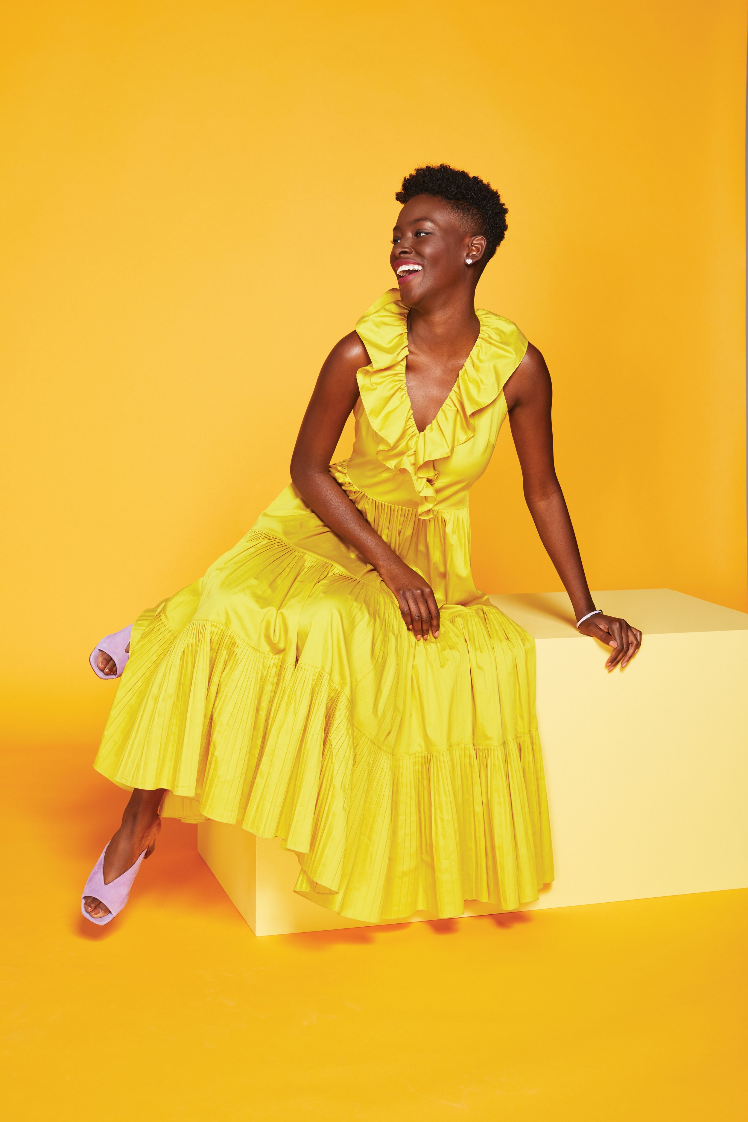 Woman in a bright yellow ruffled dress sitting on a yellow block against a yellow background, smiling and looking to her left, with short curly black hair.