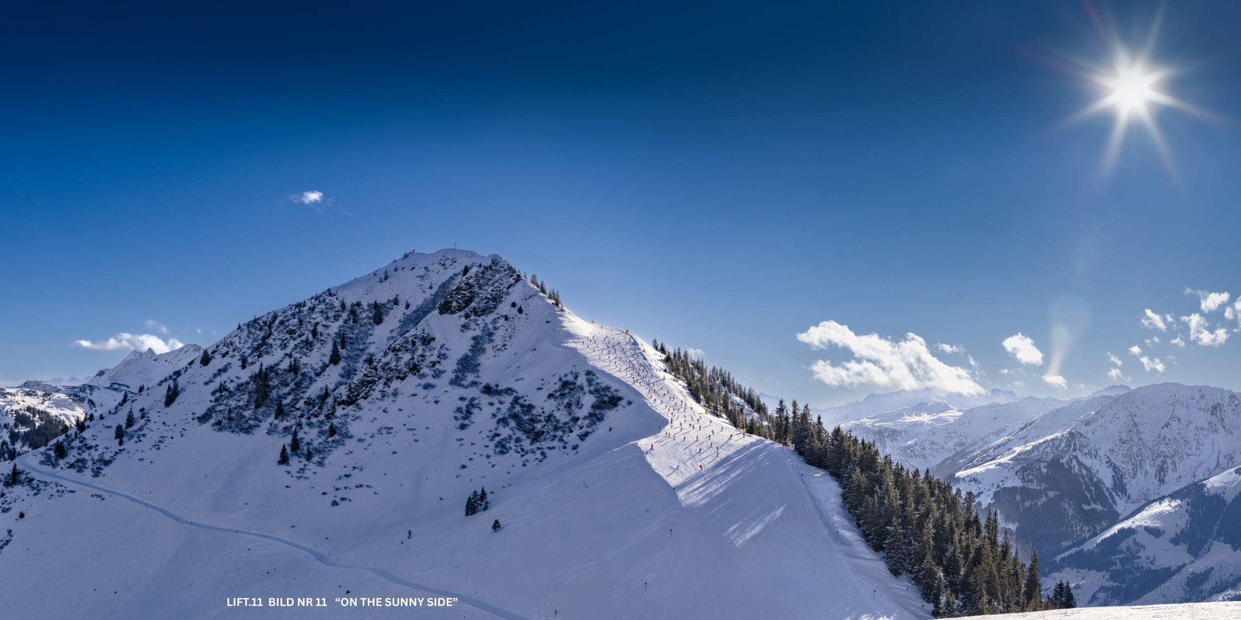 Schneebedeckte Berglandschaft bei Sonnenschein, mit Skifahrern auf einer Piste und einem blauen Himmel
