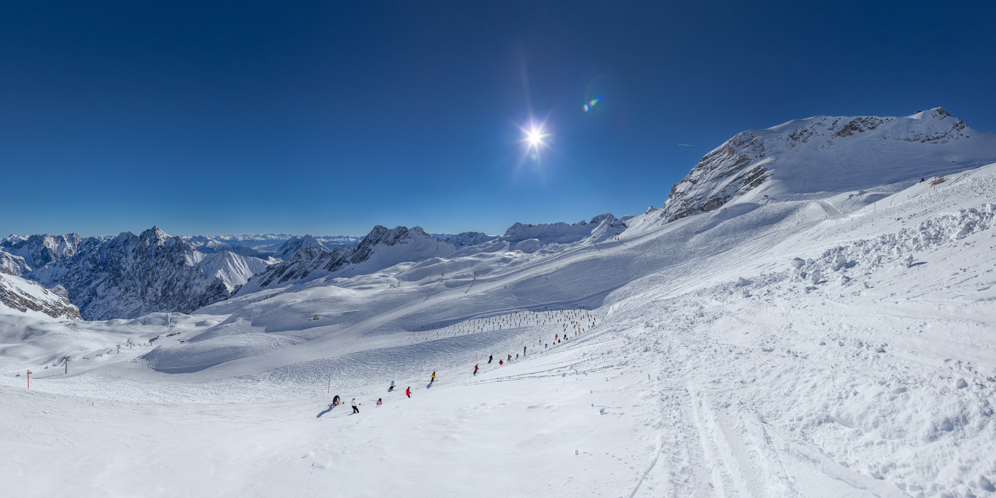 Schneebedeckte Berge bei Sonnenschein, Skifahrer auf einer Piste, klare blauer Himmel.