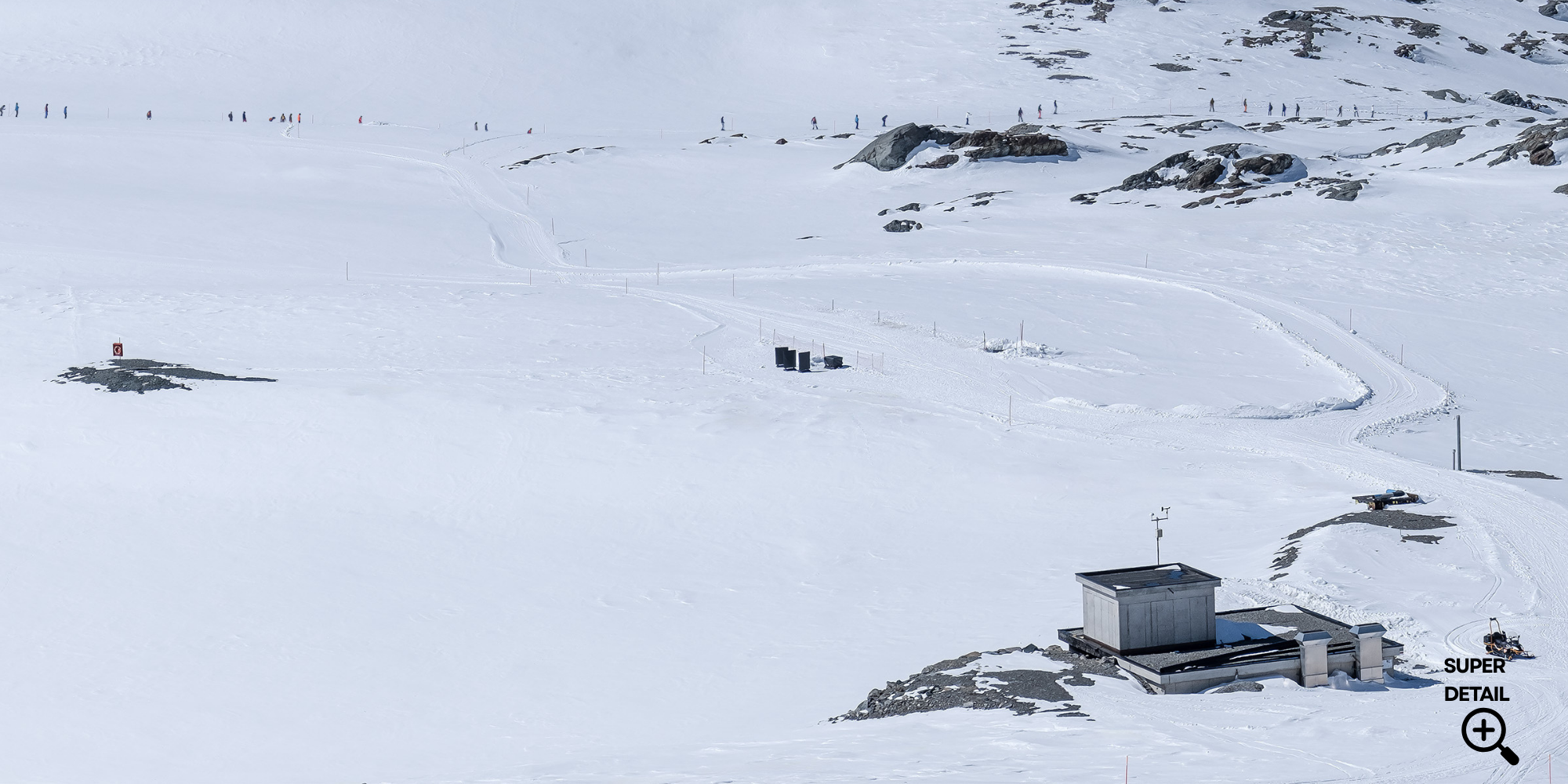 Schneebedeckte Berglandschaft mit Skifahrern in der Ferne, technische Ausrüstung und Gebäude im Vordergrund.