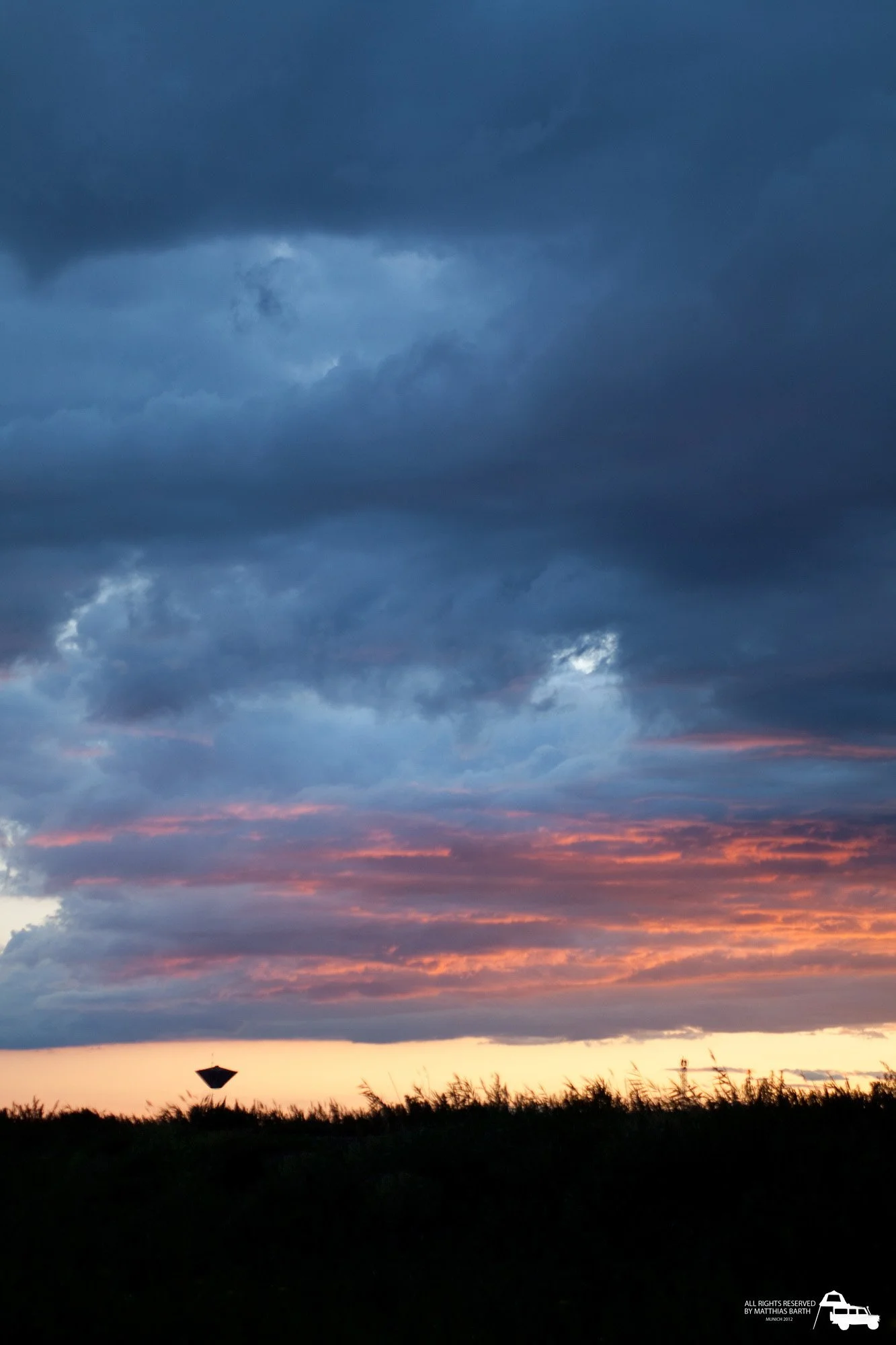 Der Himmel ist voller dunkler Wolken mit einem Farbverlauf von Blau zu Orange, während die Sonne untergeht. Im Vordergrund sind Silhouetten von Pflanzen und eine kleine, dunkle Struktur zu sehen.
