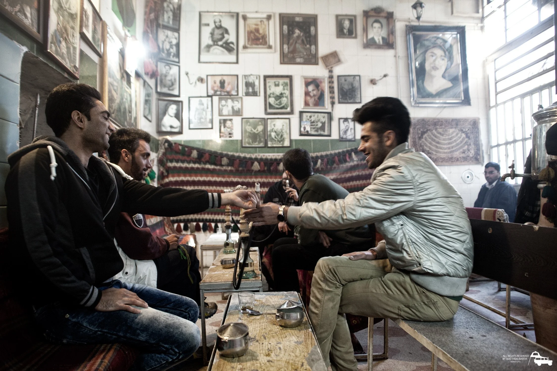 Eine Gruppe von jungen Männern sitzt in einem Café, spielt mit einer Wasserpfeife, lachen und unterhalten sich. Die Wände sind mit vielen Fotos und Gemälden geschmückt, es herrscht eine gemütliche Atmosphäre.