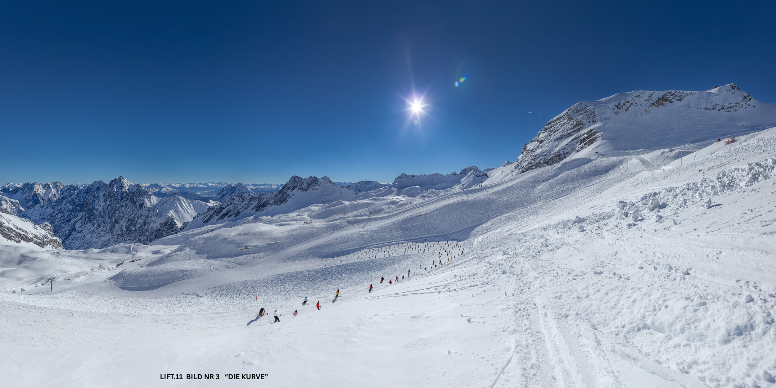 Verschneite Berge und Skifahrer auf einer Skipiste bei Sonnenschein, mit klarem blauen Himmel.