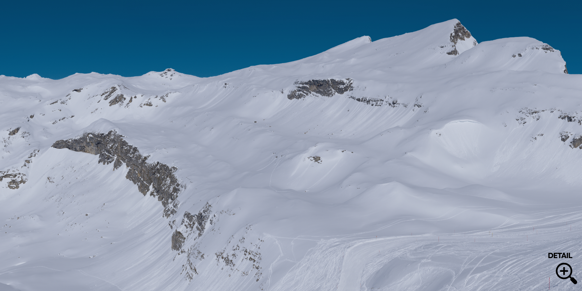 Schneebedeckte Berge unter blauem Himmel, Skispuren im Schnee im Vordergrund.