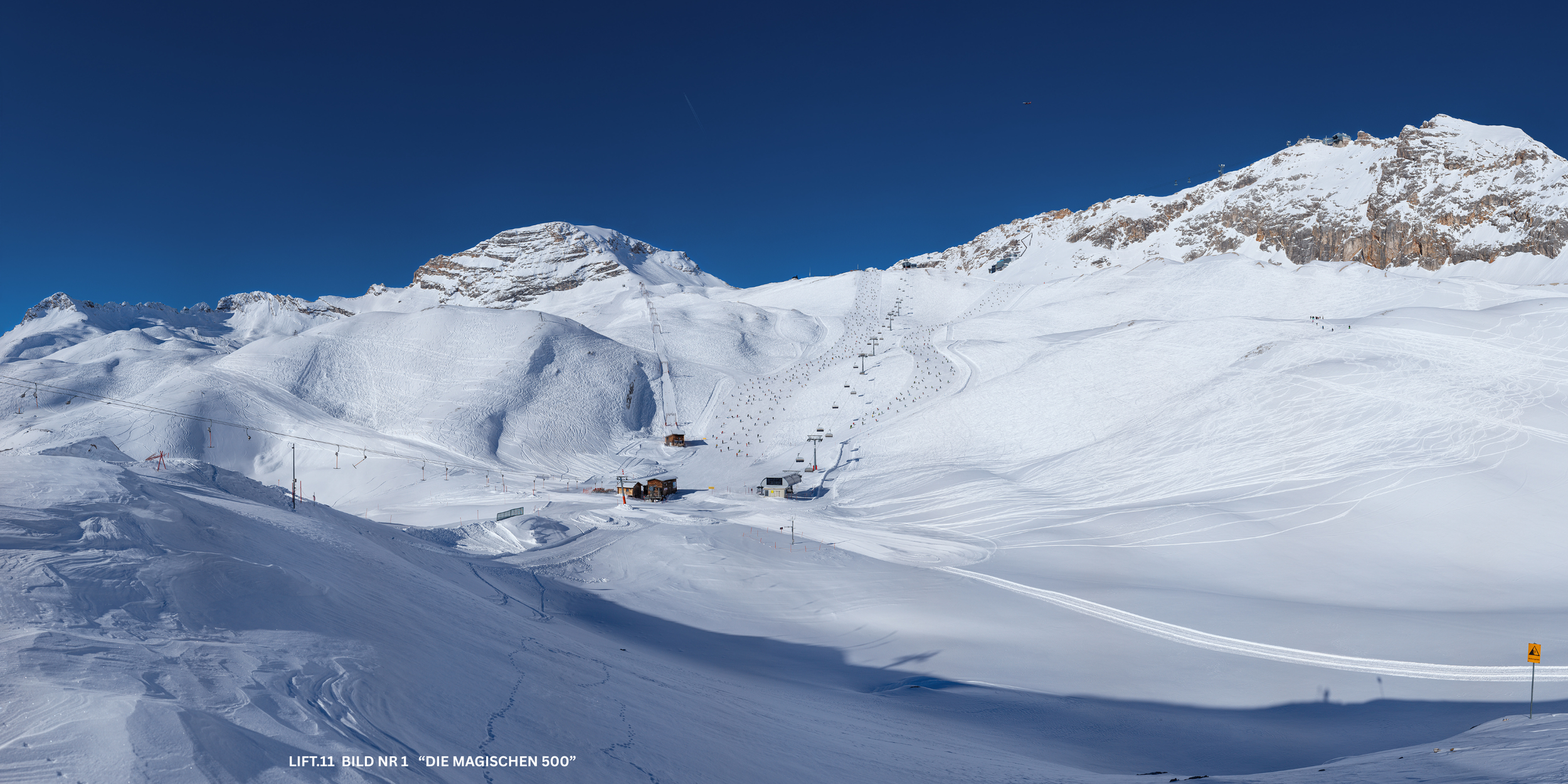 Verschneite Skipiste in den Bergen mit Skiliften und einem kleinen Gebäude, unter einem klaren blauen Himmel.