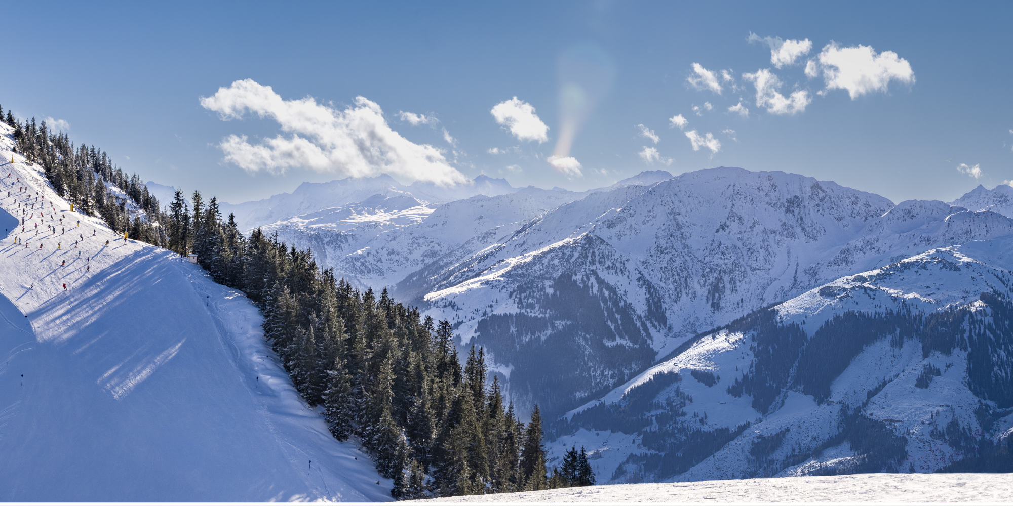 Gelisene Pisten mit Skifahrern in den Bergen, umgeben von schneebedeckten Gipfeln und Tannen im klaren blauen Himmel.