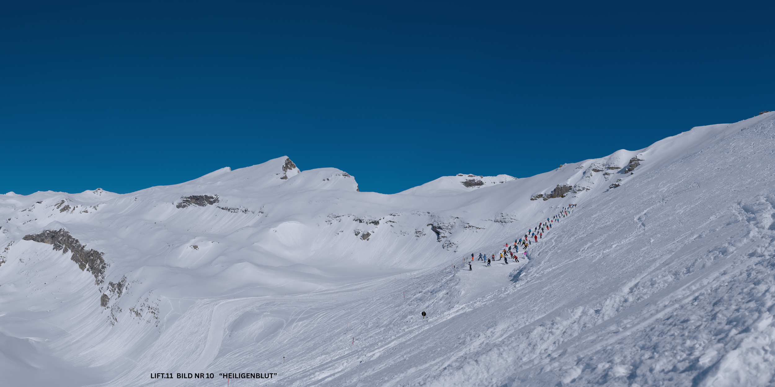 Schneebedeckte Berge mit Skifahrern in einer langen Reihe, die einen Hang hinauffahren.