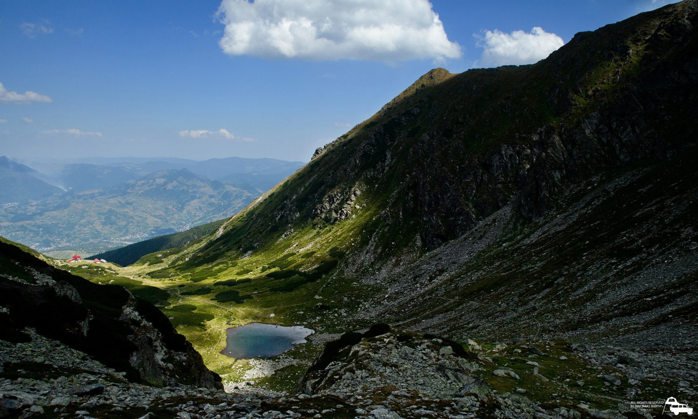 Bergsee in einem Tal mit grünen Wiesen, umgeben von steilen, felsigen Bergwänden unter einem blauen Himmel mit Wolken.