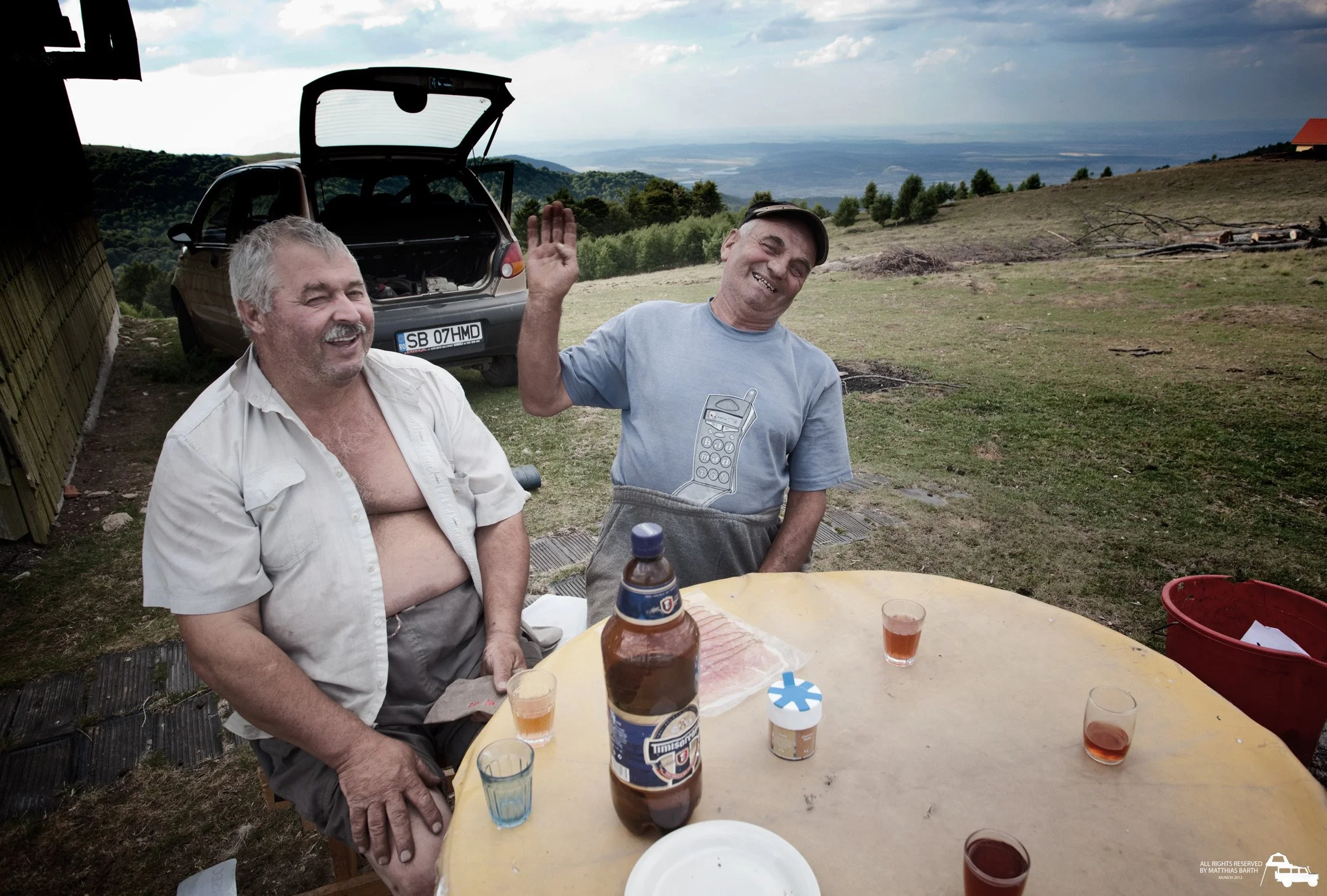 Zwei Männer sitzen an einem runden Tisch im Freien, trinken und lachen. Im Hintergrund ein Auto mit offenem Kofferraum und eine hügelige Landschaft mit Bäumen und Gebäuden. Die Männer wirken entspannt und fröhlich.