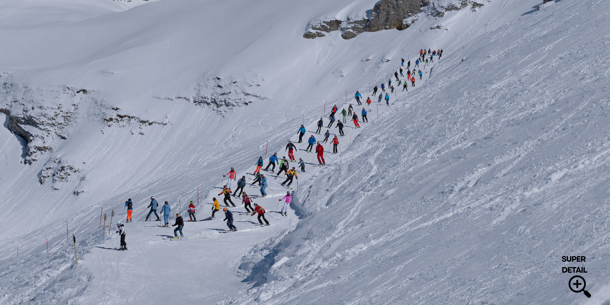Viele Skifahrer in bunten Kleidung auf einer verschneiten Skipiste im Gebirge, einige auf dem Boden, andere beim Skifahren, mit Felsen im Hintergrund.