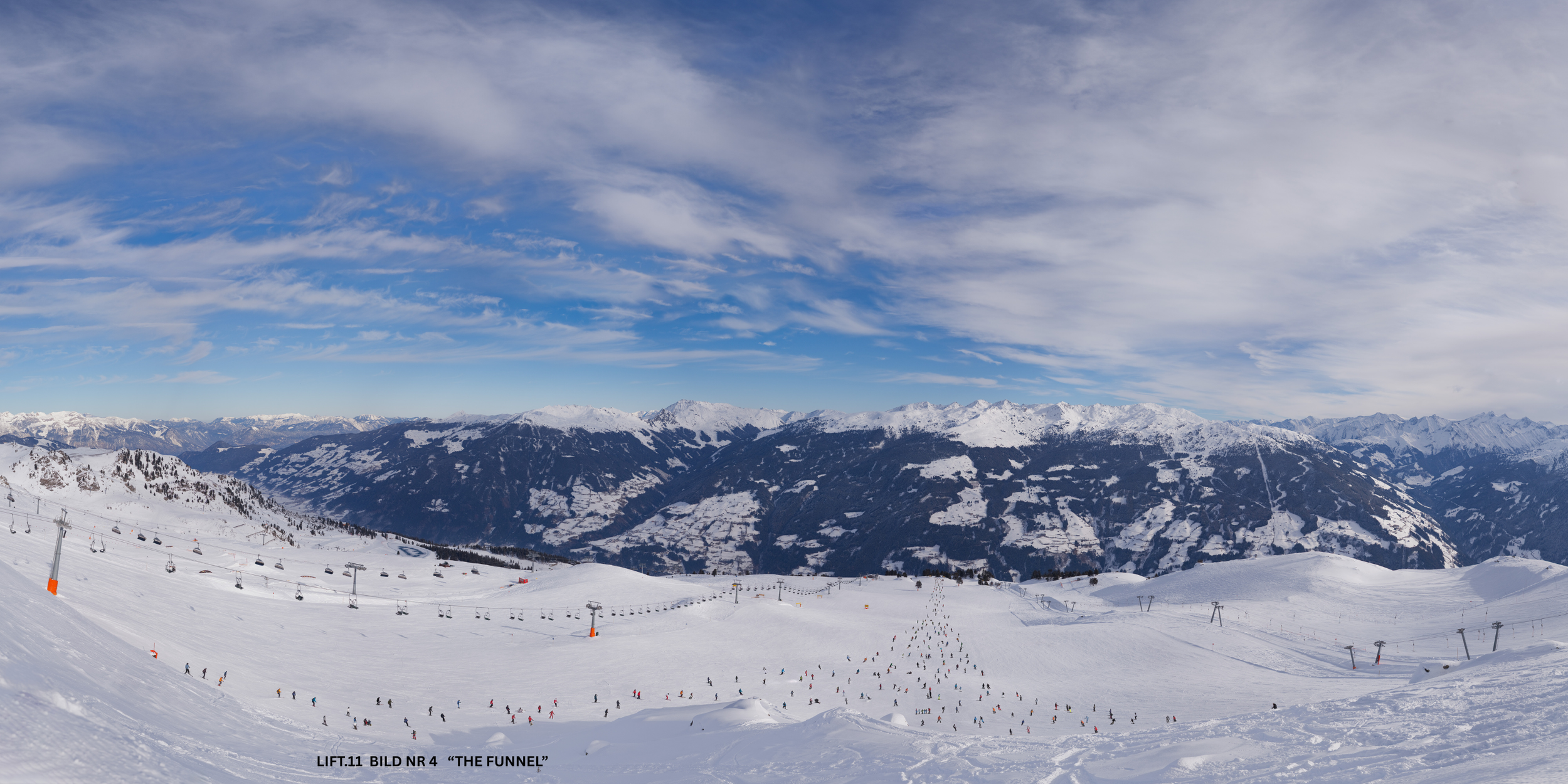 Bergpanorama im Schnee mit Skilift, Skifahrern und schneebedeckten Bergen im Hintergrund unter einem bewölkten Himmel.
