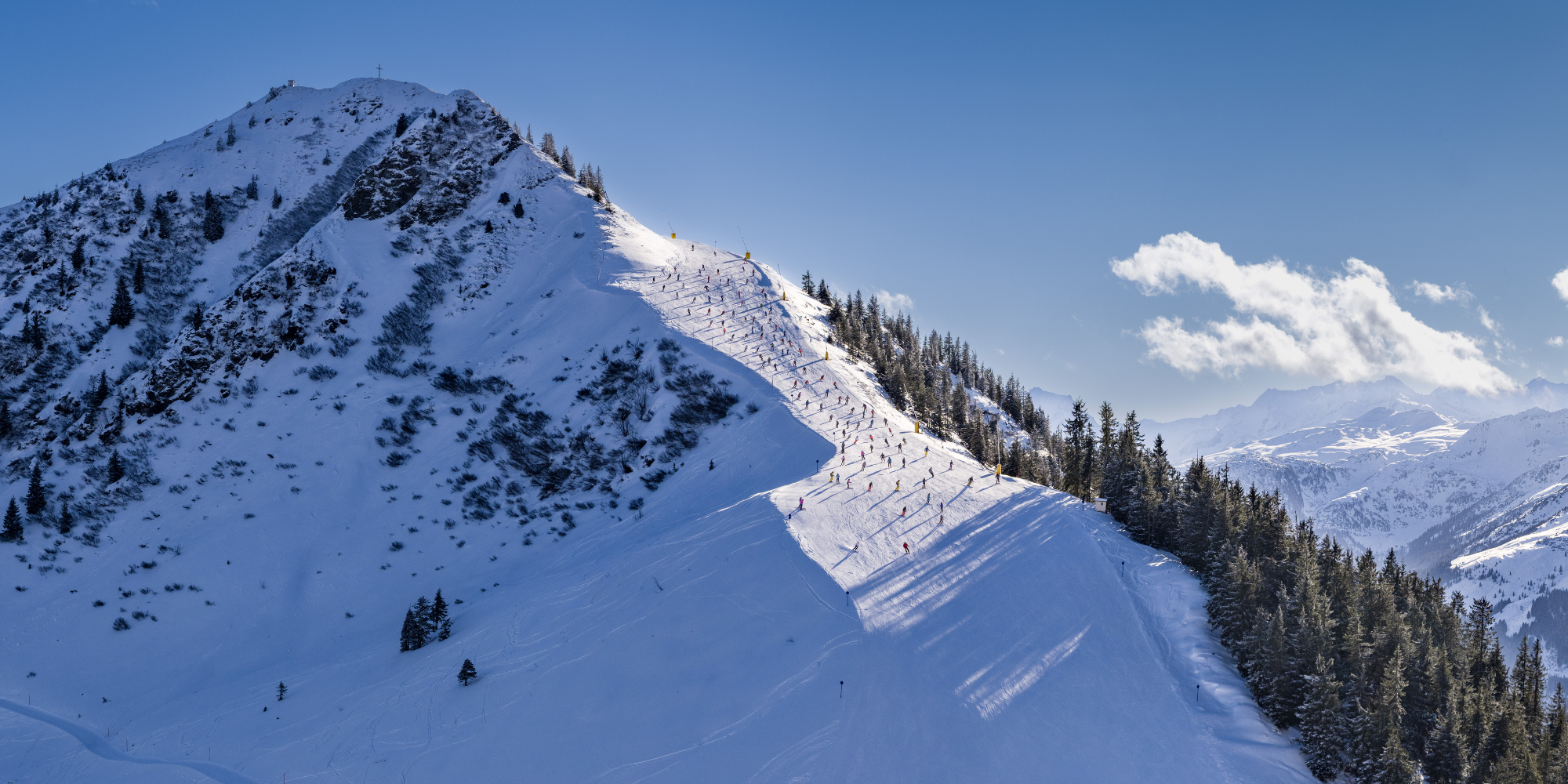Berg mit Skifahrern auf einer Piste, Schneelandschaft und Wolken im Hintergrund.