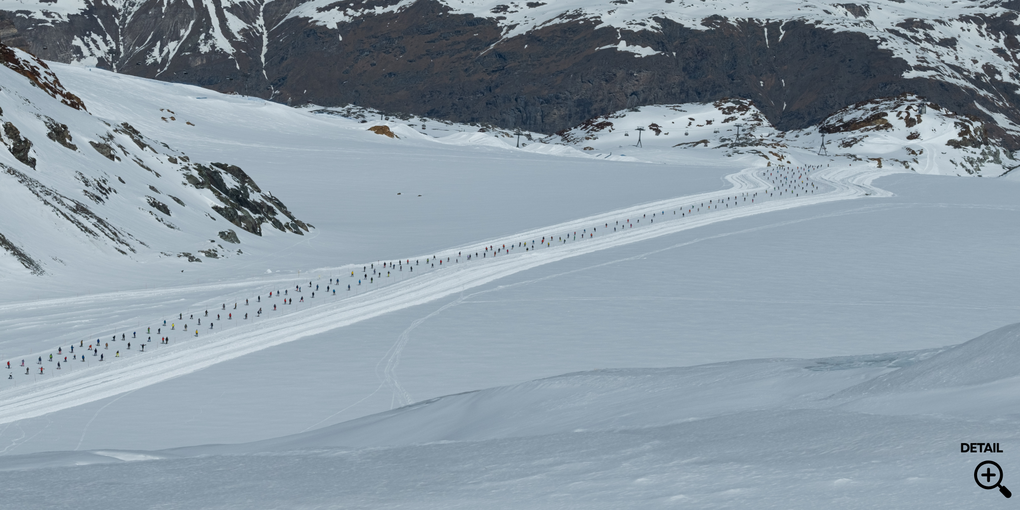 Schneeweiße Hochgebirgslandschaft mit einer langen Schneespur, auf der eine große Gruppe von Skifahrern in einer Reihe läuft, vor Bergen mit Schneeflecken und Skiliften im Hintergrund.