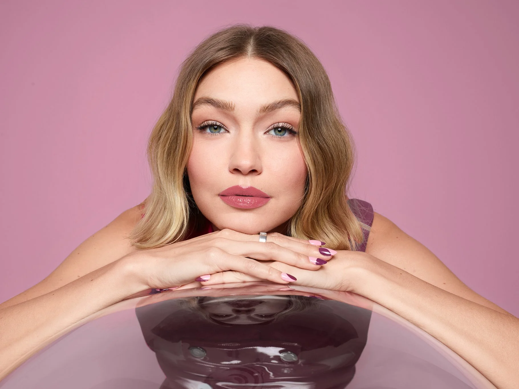 A woman with blonde hair and pink lipstick leaning on her hands, with a mirror reflecting her face, against a pink background.