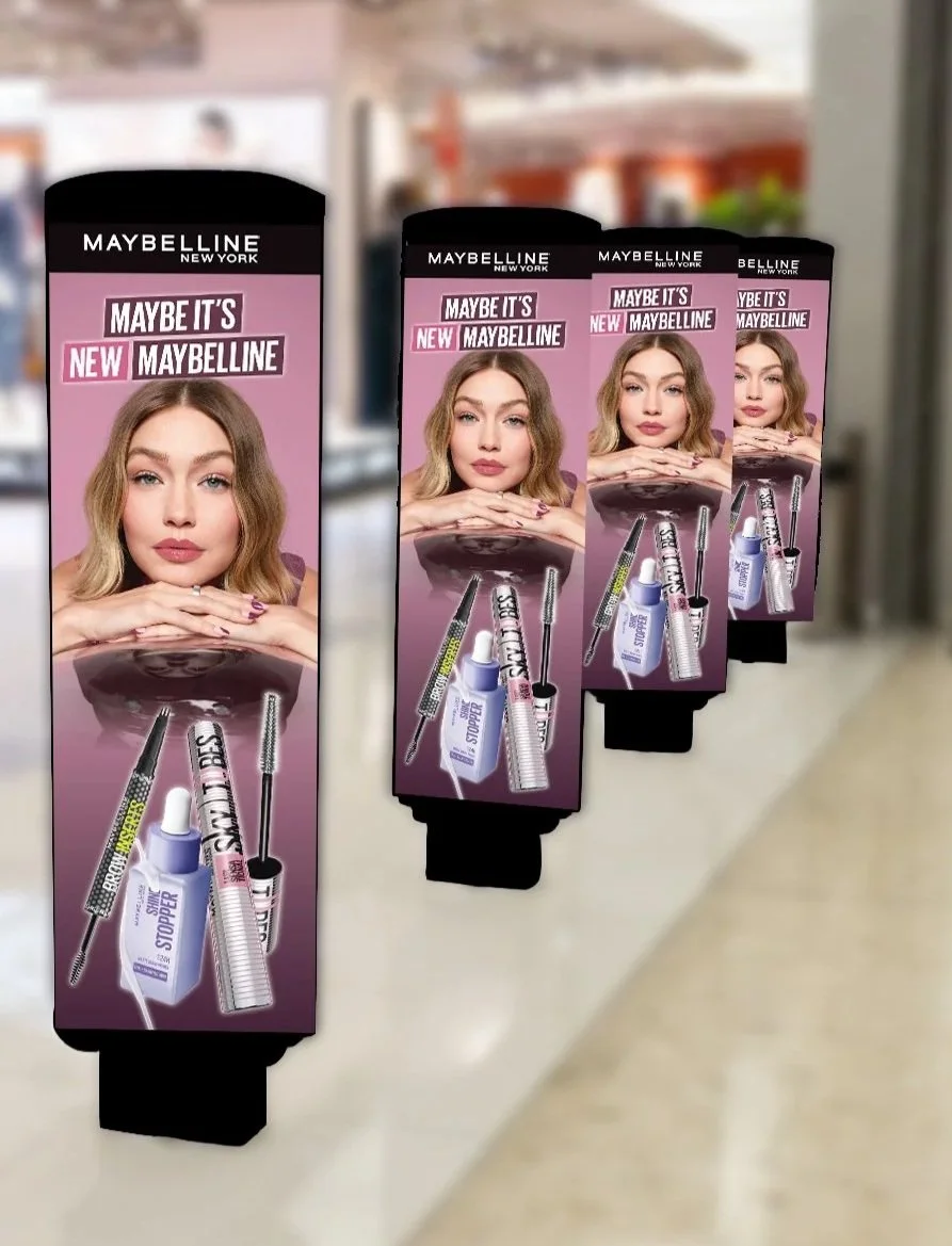 Three makeup advertisement signs in a store, featuring a young woman with blonde hair and neutral makeup, promoting Maybelline products. Each sign displays images of various Maybelline makeup items including mascara, eyeliner, and setting spray.