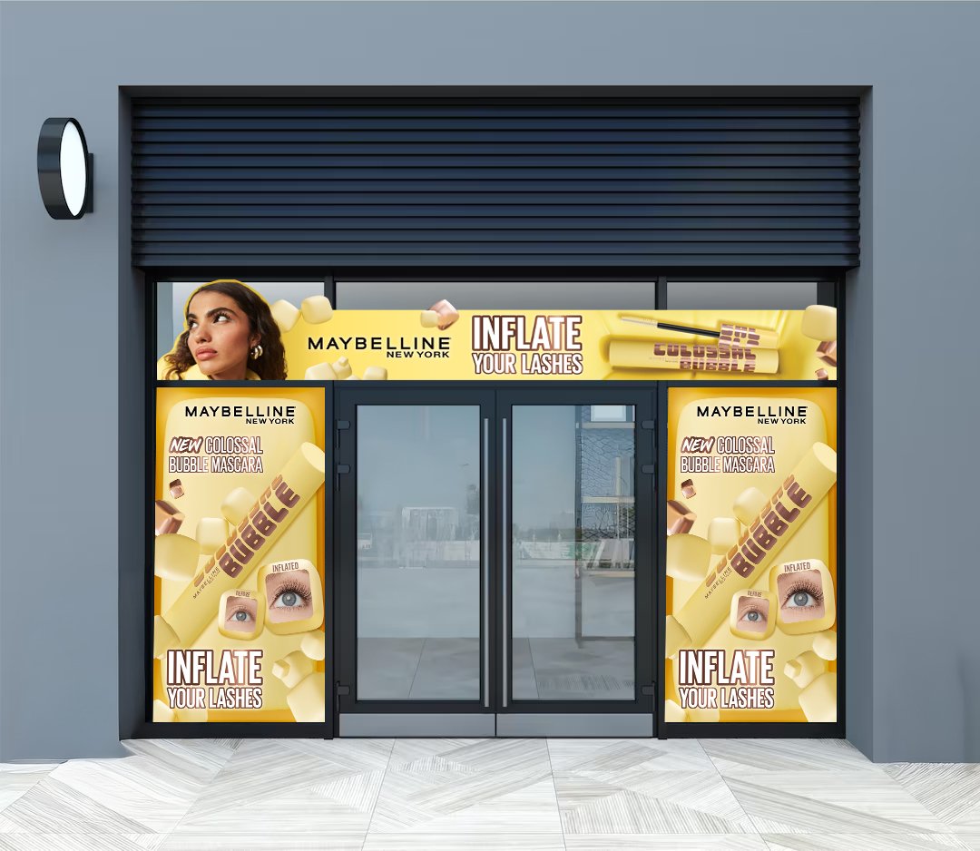 storefront advertisement for Maybelline New York promoting a new colossal bubble mascara that claims to inflate lashes, with colorful graphics and close-up of an eye with eyelash enhancement.