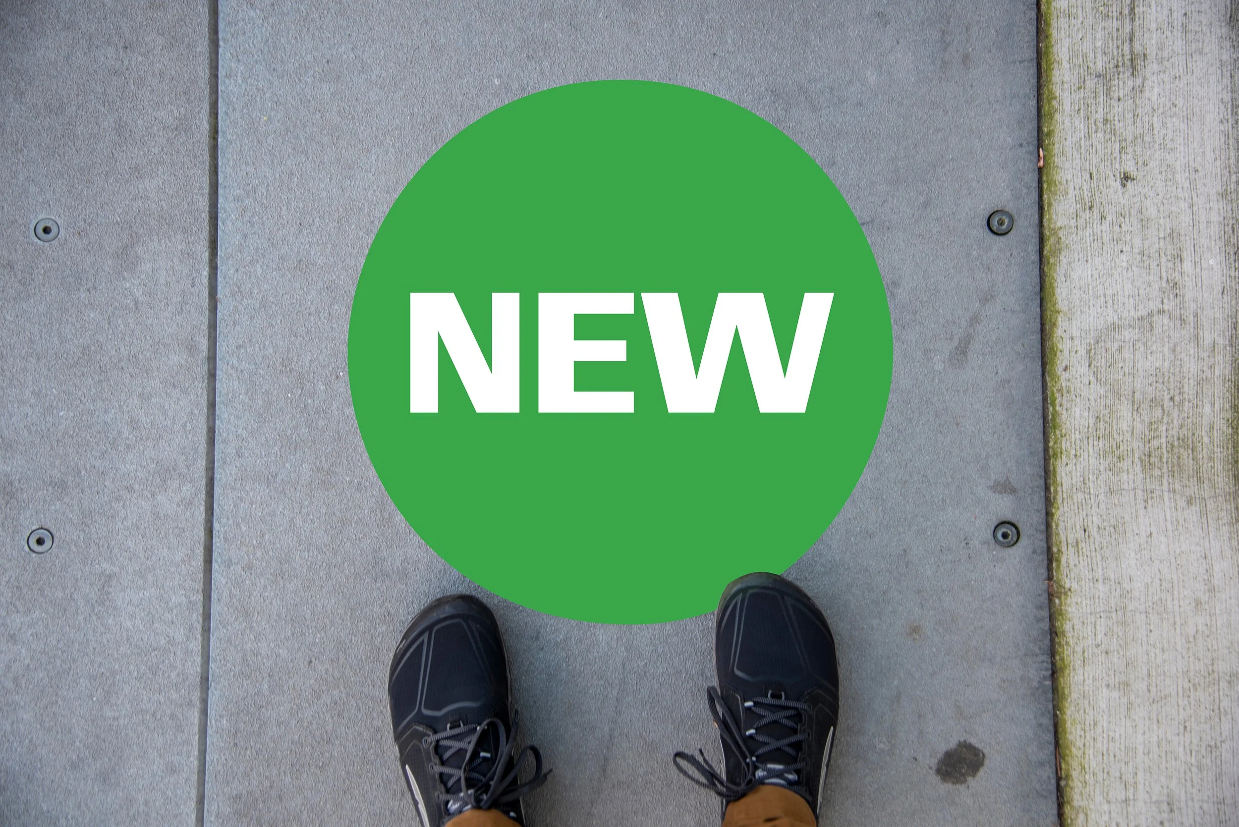 A person standing on a sidewalk looking down at a green circular sticker on the ground that says 'NEW' in white letters, with black shoes and brown pants visible.