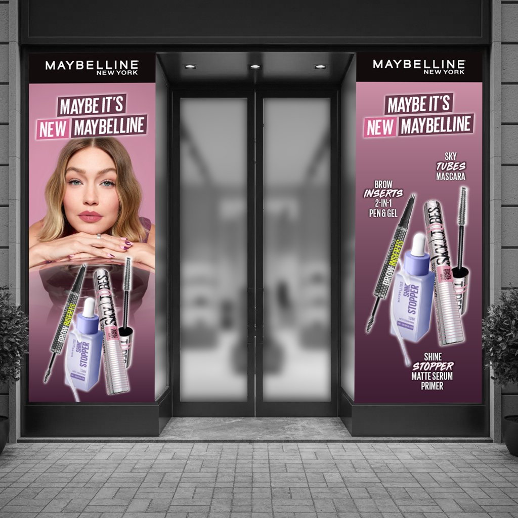 Two large vertical banners on either side of a glass door feature Maybelline New York makeup products and a model with wavy blonde hair on a pink background. The left banner showcases a close-up of the model's face with her hands under her chin, and displays products including Sky Tubes mascara, Brow Insert 2-in-1 pen and gel, and Shine Stopper matte serum primer. The right banner lists the same products with descriptions: Sky Tubes mascara, Brow Insert 2-in-1 pen and gel, and Shine Stopper matte serum primer.
