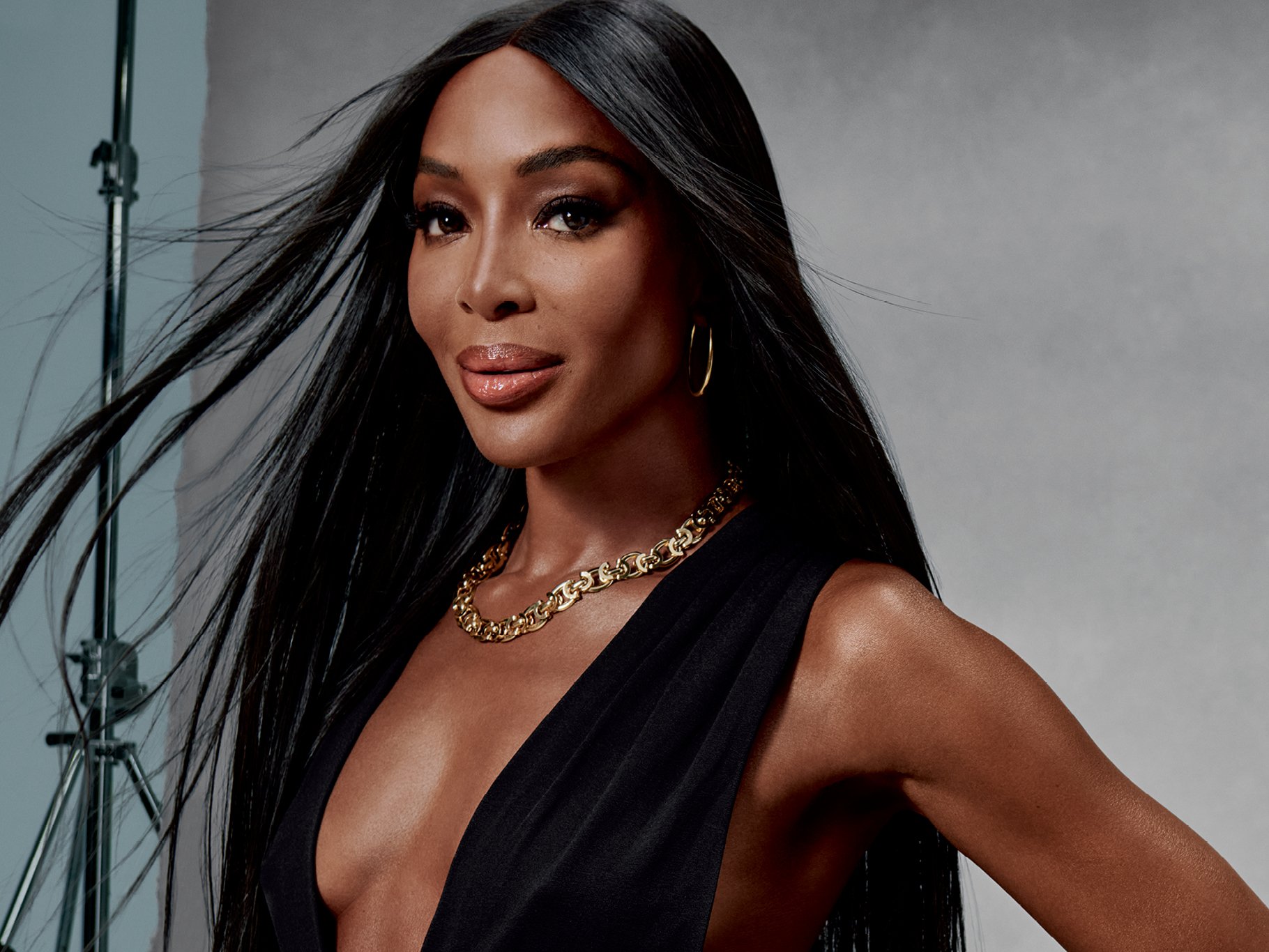 Naomi Campbell of the Maybelline 'Like an Icon' Campaign, wearing gold jewelry including a chain necklace and hoop earrings, dressed in a sleeveless black top, posing in front of a gray background, with professional lighting equipment visible.