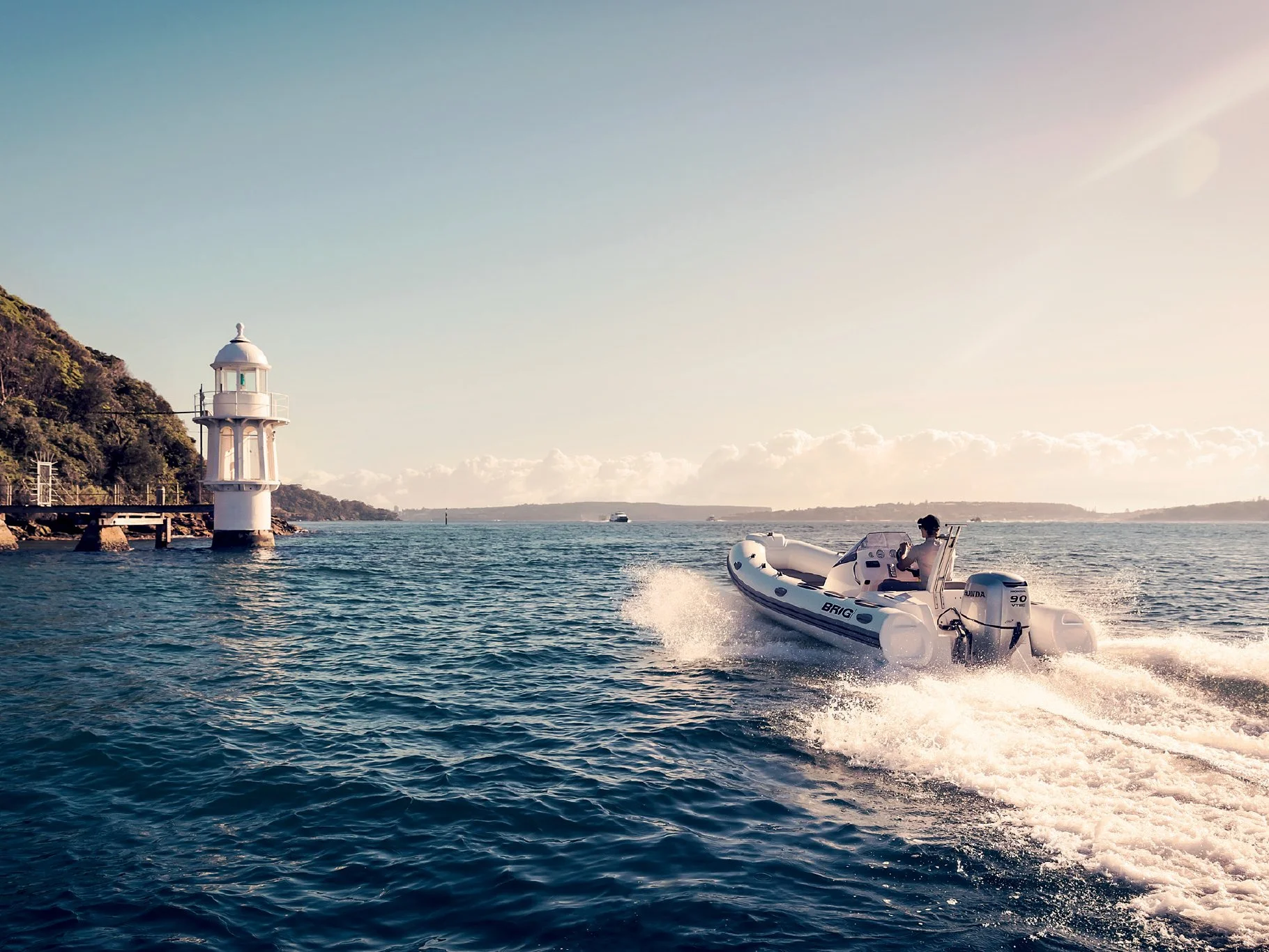 A person driving a white BRIG boat on open water near a lighthouse on a shoreline with a hill and cloudy sky in the background.