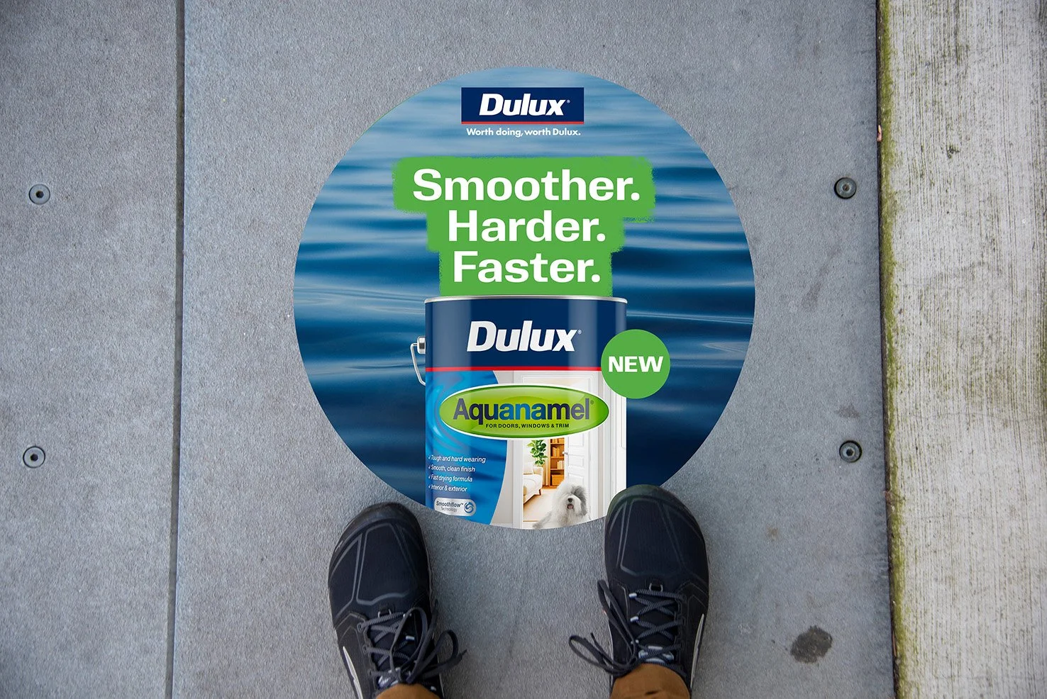 Footwear standing on ground over a circular advertisement for Dulux Aquanamel paint, featuring the tagline 'Smoother. Harder. Faster.' with water background and a paint can image.