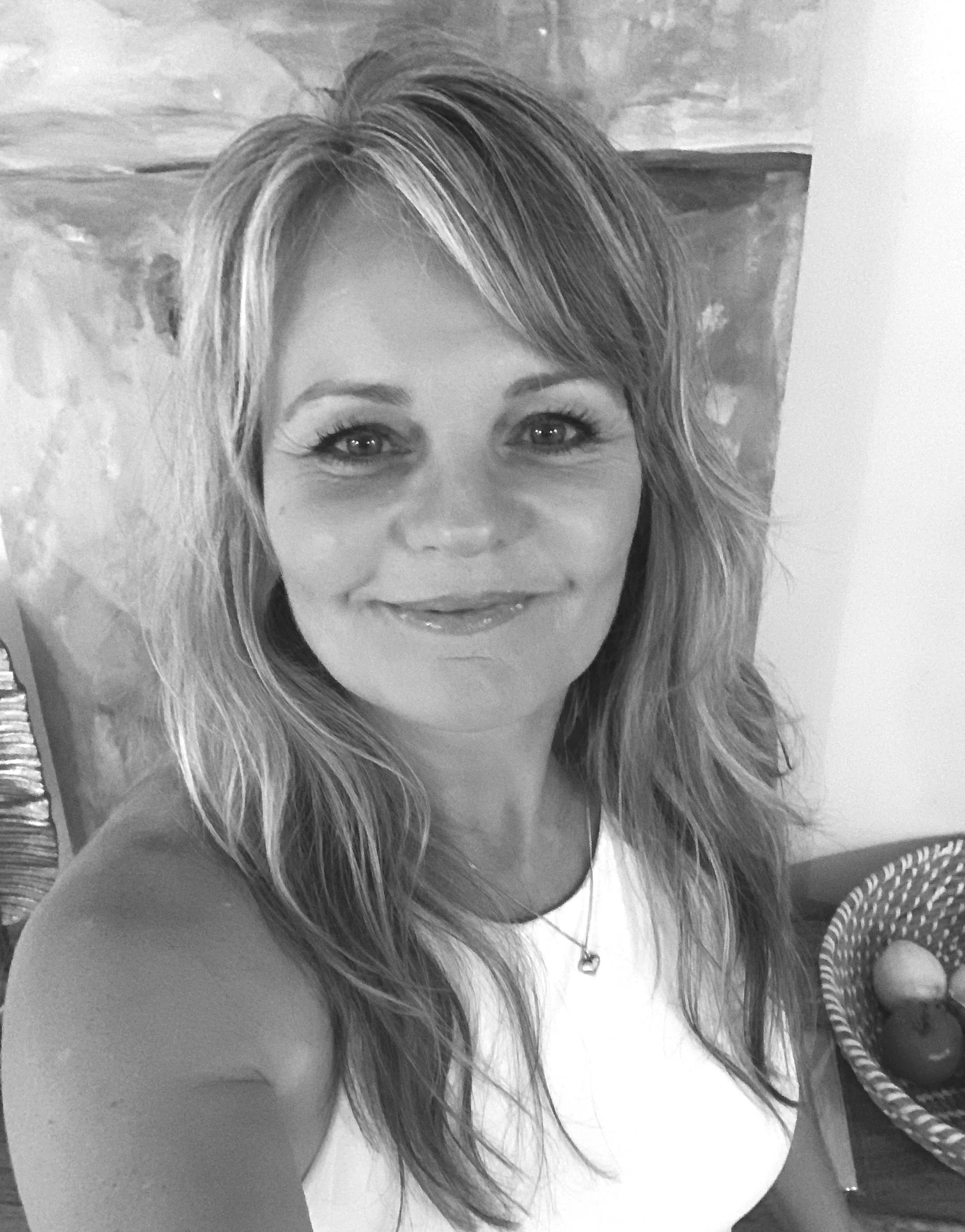 Black and white photo of a woman with long, wavy hair and a big smile, wearing a necklace with a heart pendant.