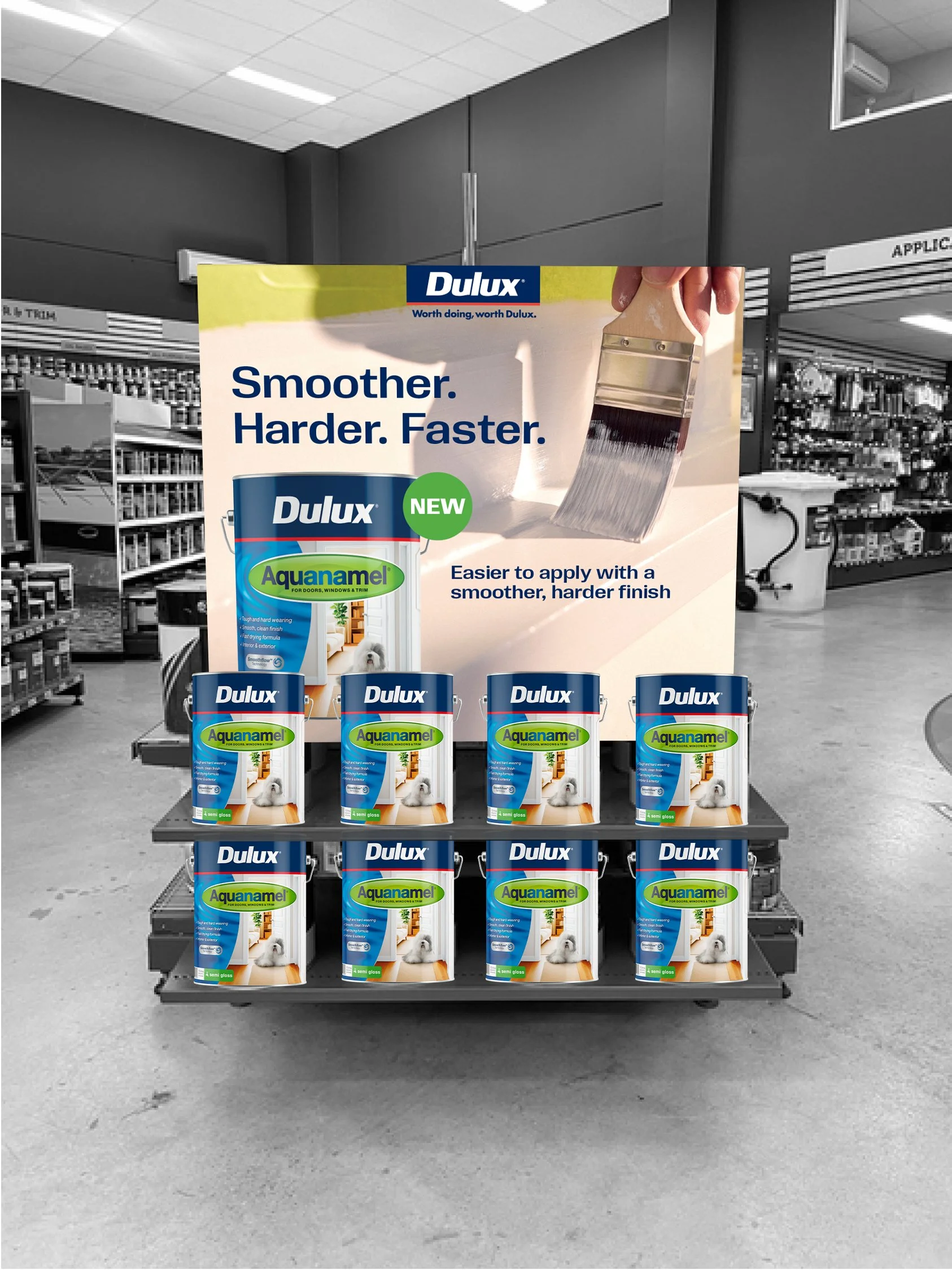 Display in a store aisle showing multiple cans of Dulux Aquanamel semi-gloss paint arranged on a metal shelf with a promotional poster behind them. The poster features a person applying paint with a brush, and text that reads 'Smoother. Harder. Faster.' and 'Easier to apply with a smoother, harder finish.' The store has a gray interior with shelves of various products in the background.