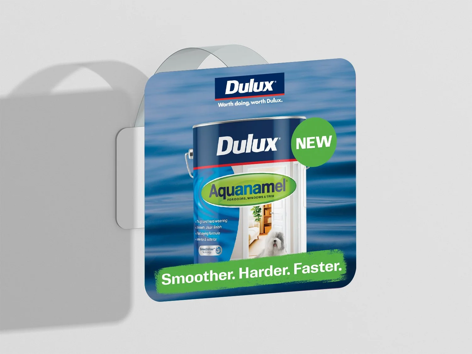 A wall-mounted air freshener dispenser with a label for Dulux Aquanamel paint, showing a can of paint and a window with a view to a room with a dog inside, and a green circle indicating it is new, with a green slogan at the bottom that says 'Smoother. Harder. Faster.'.