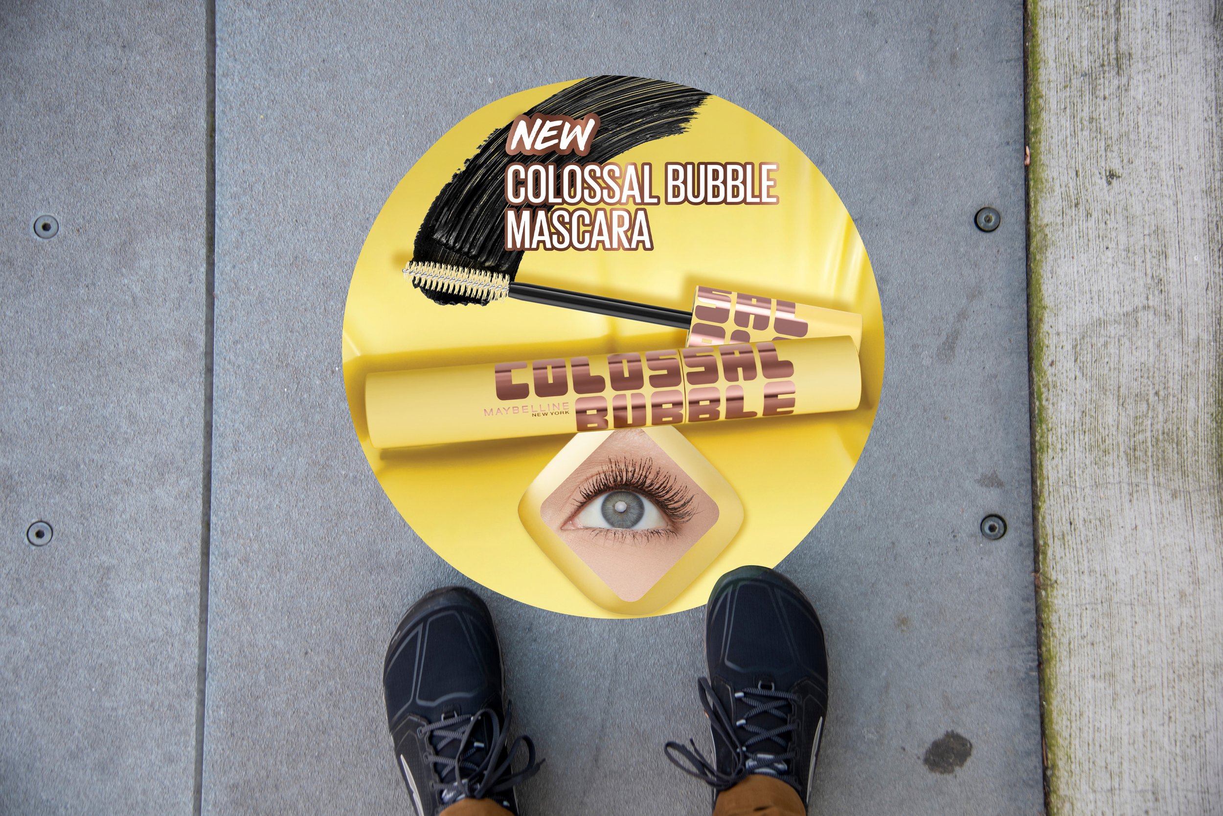 View of a person standing on a sidewalk, wearing black sneakers and brown pants, looking down at advertising materials for Maybelline New York's new Colossal Bubble mascara, including a tube and a digital image of an eye with long eyelashes