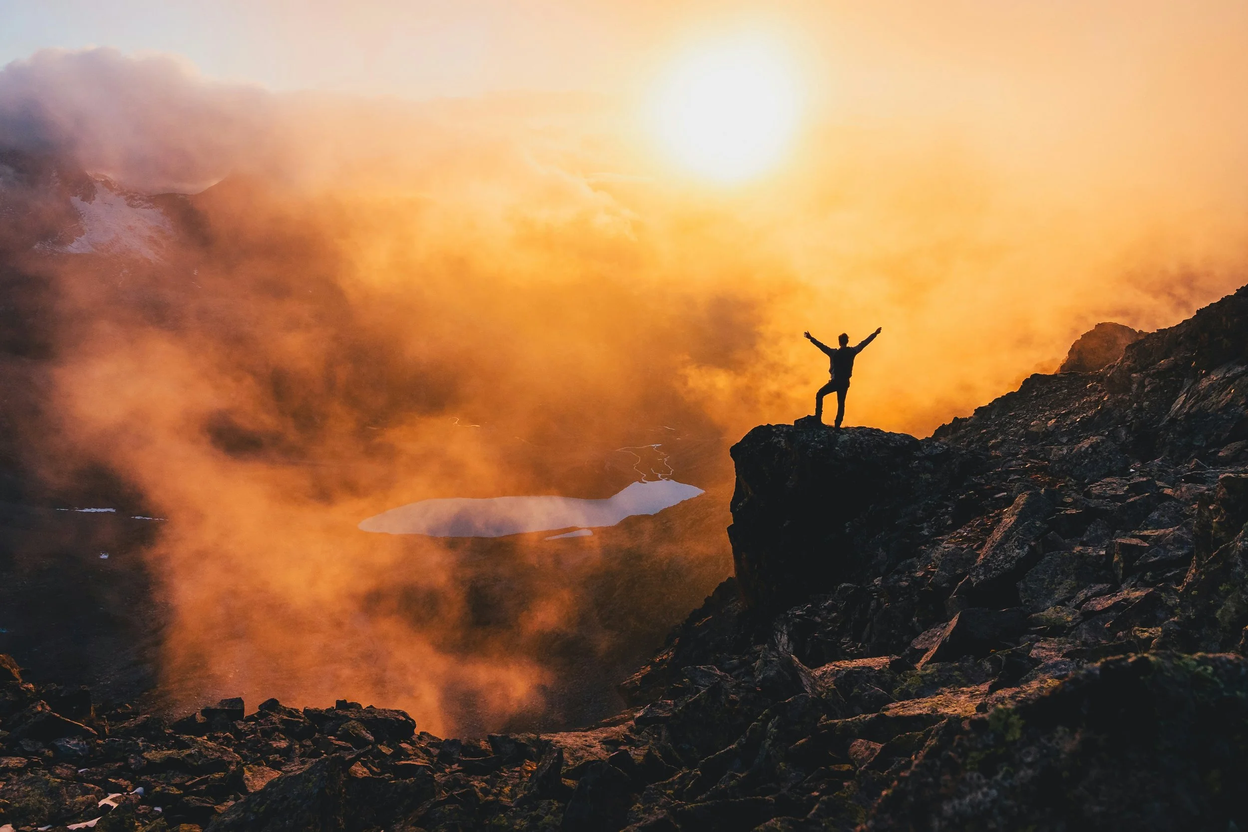 Silhouette of a person standing on rocky terrain with arms raised, overlooking a landscape with clouds and a bright sun in the sky at sunset.