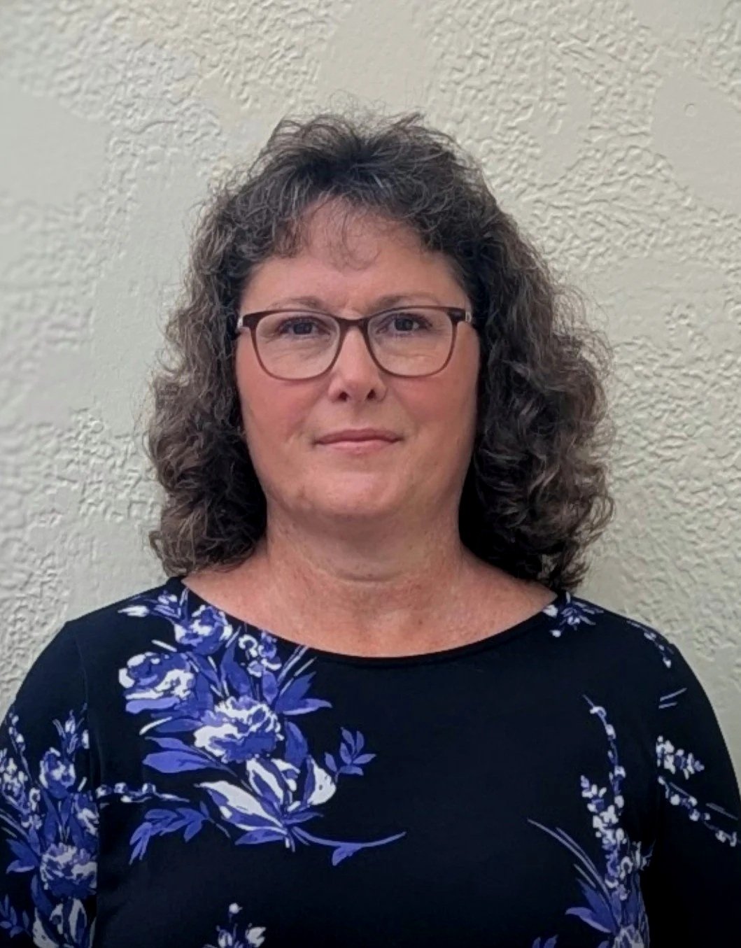 A woman with curly gray hair and glasses, wearing a black shirt with a blue flower pattern, standing against a textured beige wall.