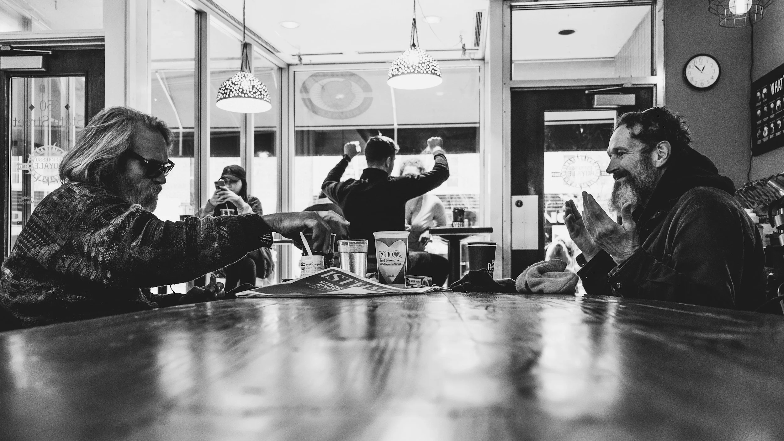 Deux hommes âgés assis en face l'un de l'autre dans un café, échangeant un sourire, avec un groupe de personnes en arrière-plan, dont une en train de prendre une photo et une autre levant les bras.