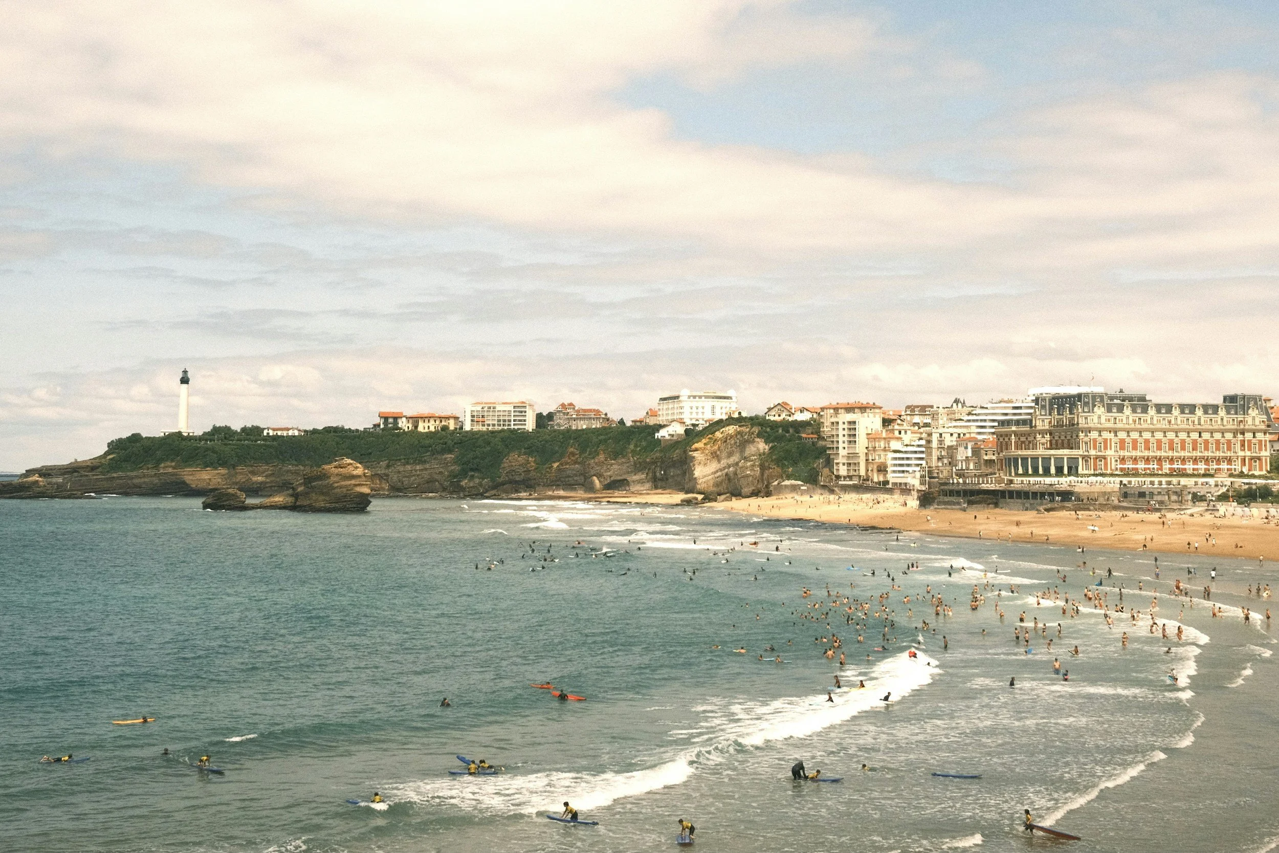 Plage animée avec de nombreux surfeurs dans l'eau et des gens sur le sable, côte avec falaises et un phare en arrière-plan sous un ciel partiellement nuageux.