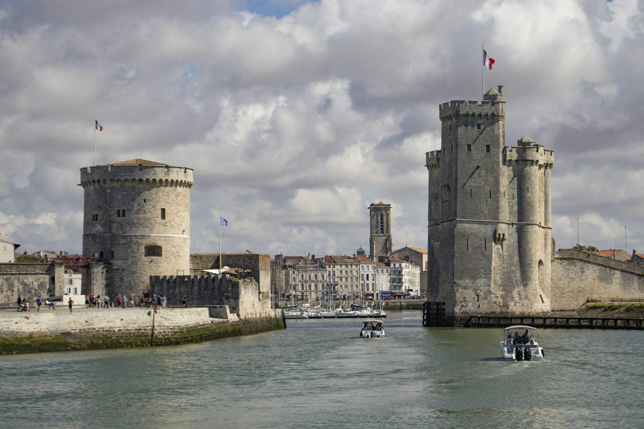 Deux tours médiévales en pierre surplombant un port avec bateaux, sous un ciel nuageux.