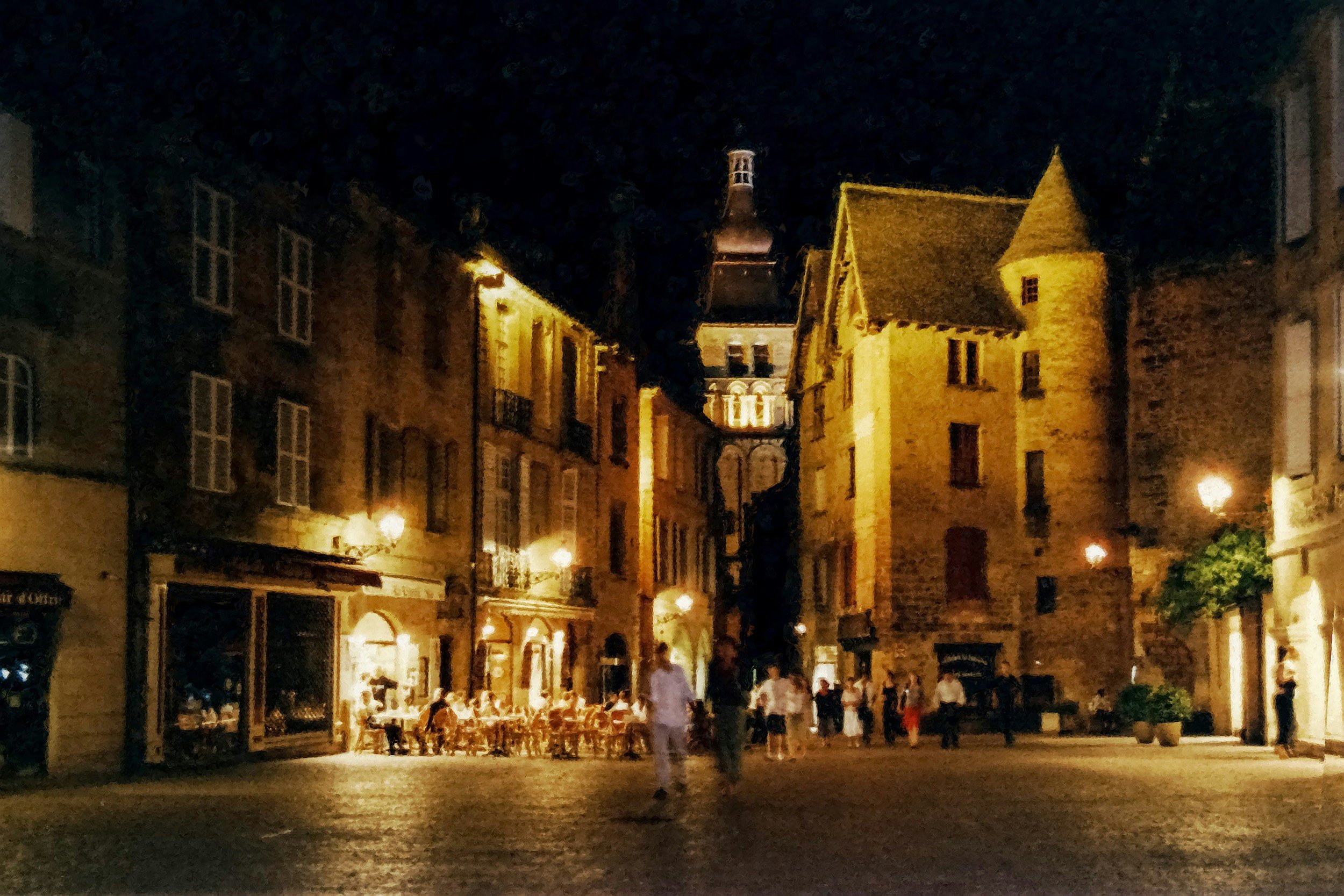 Rue pavée en soirée avec des bâtiments anciens bien éclairés, quelques personnes marchant, une terrasse de café avec des chaises, et un clocher en arrière-plan sous un ciel nocturne.