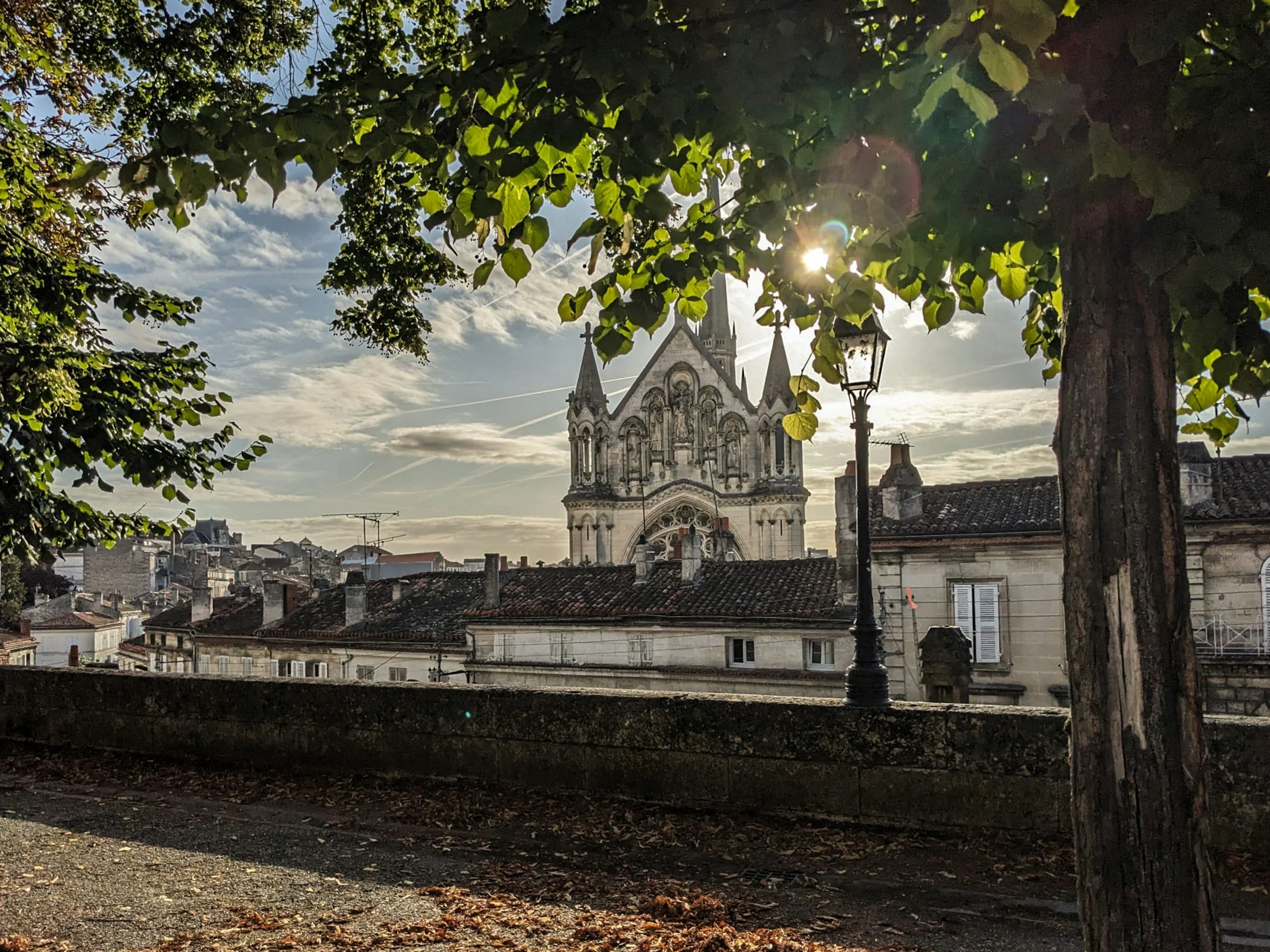 Vue de l'église gothique en arrière-plan avec des arbres et une lumière du soleil filtrant à travers les feuilles, toit en tuiles des bâtiments en premier plan.