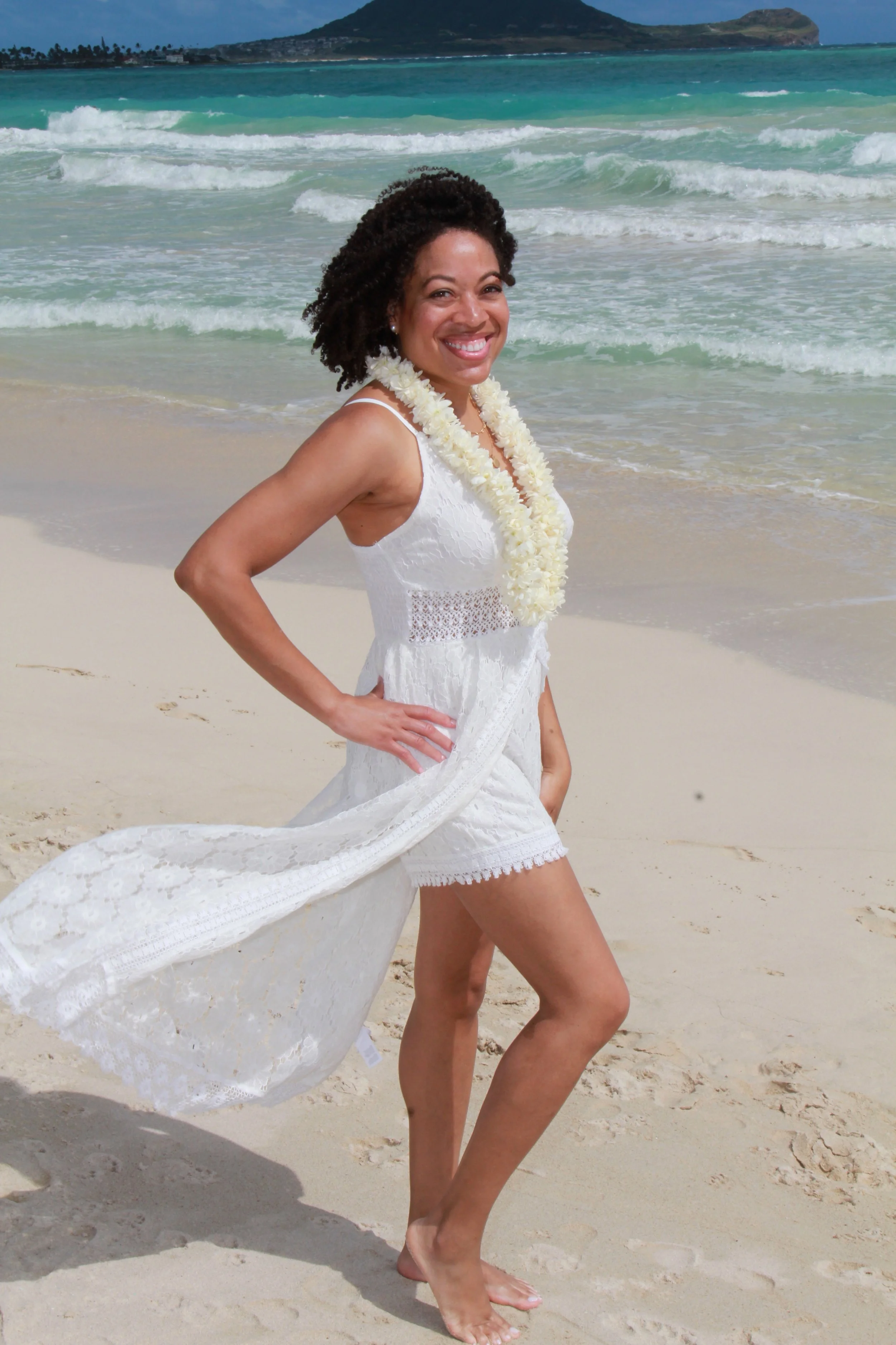 Woman in a white lace dress and a flower lei standing barefoot on a sandy beach with ocean waves and mountains in the background.