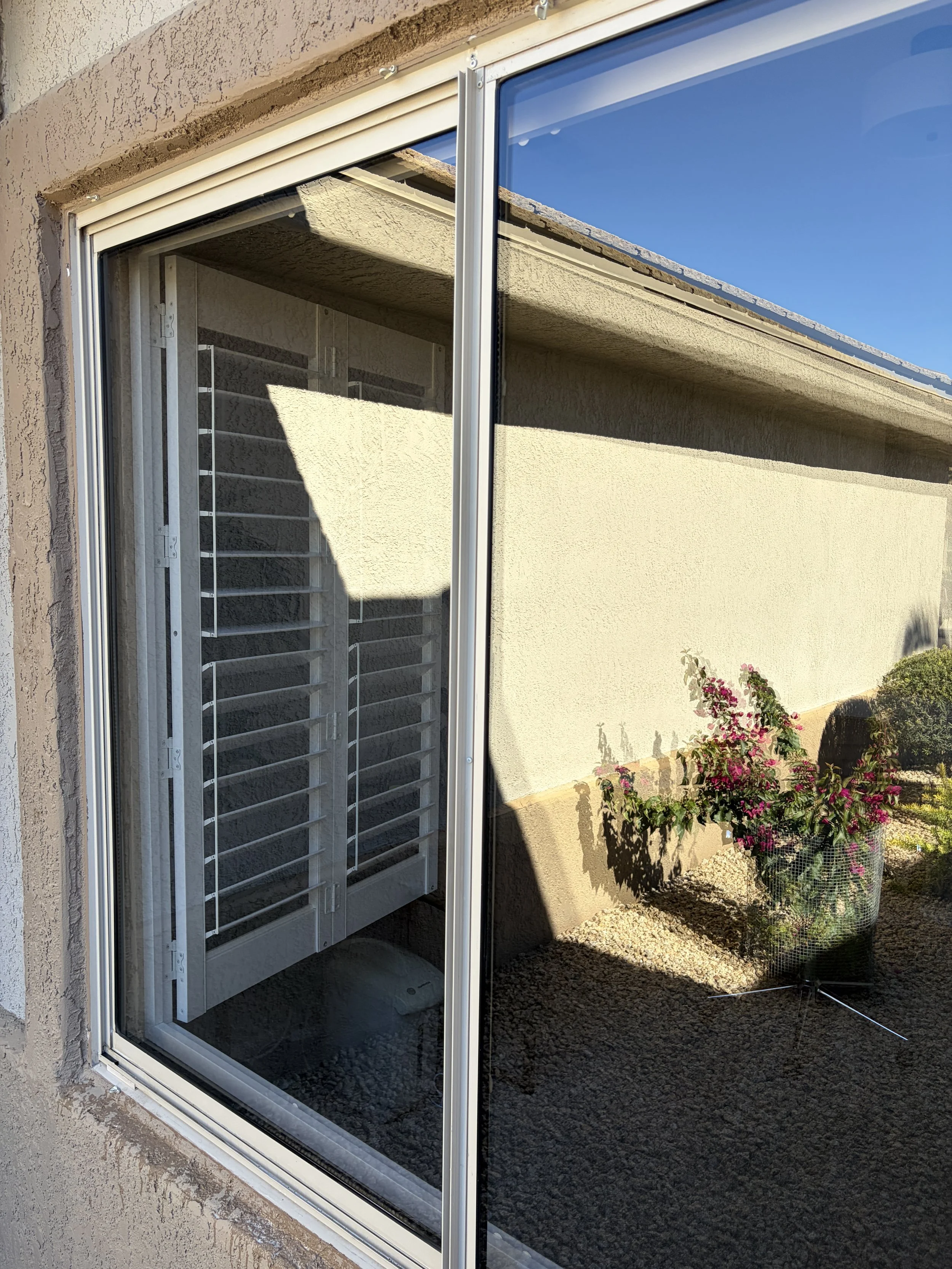 Photo of a sliding glass door reflecting a clear blue sky, a potted bush with pink flowers outside, and a building's overhang with a shadow cast on the wall. Windows cleaned by Corona Power Cleaning.