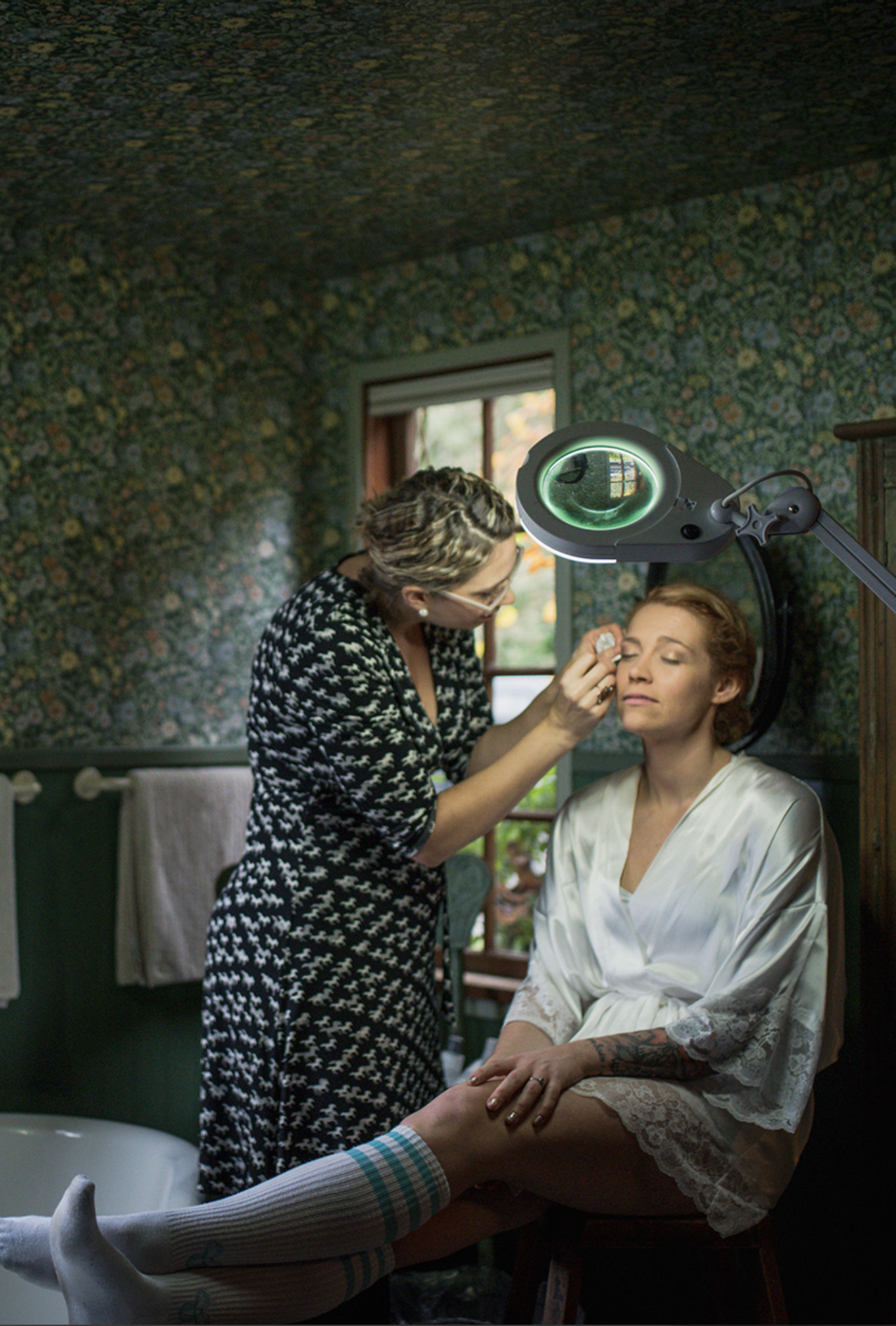 A woman in a white silk robe getting her makeup done by a makeup artist in a room with floral wallpaper
