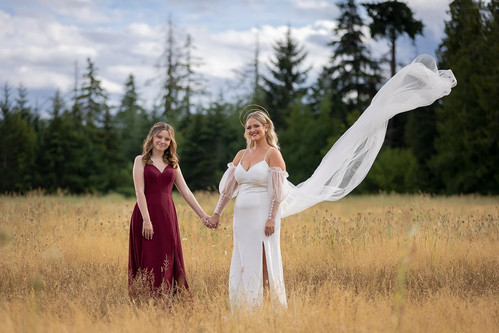 A bride and a maid of honor holding hands in a field, with trees and a cloudy sky in the background. The bride is wearing a white wedding dress with sheer sleeves and a flowing veil, while the maid of honor is in a burgundy dress.