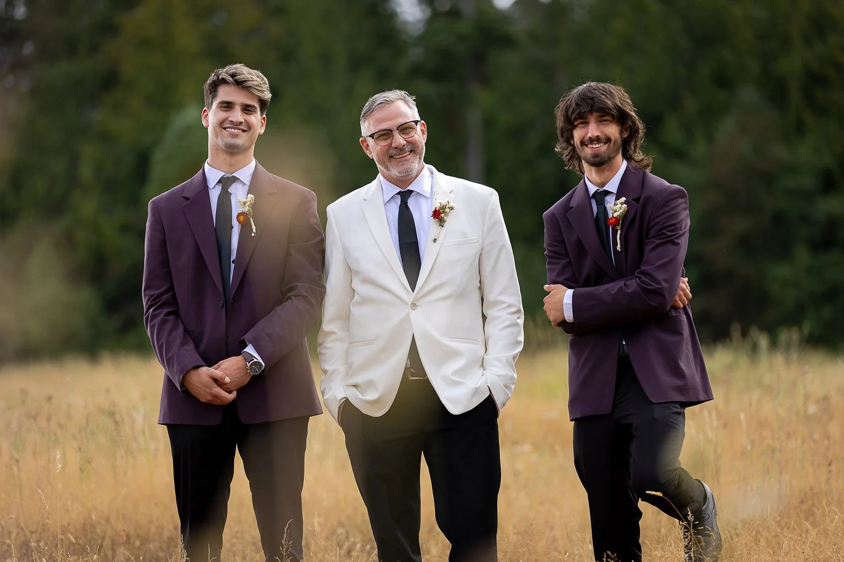 Three men in suits stand and smile outdoors in a grassy field with trees in the background. The man in the middle wears a white blazer, while the other two wear purple blazers.