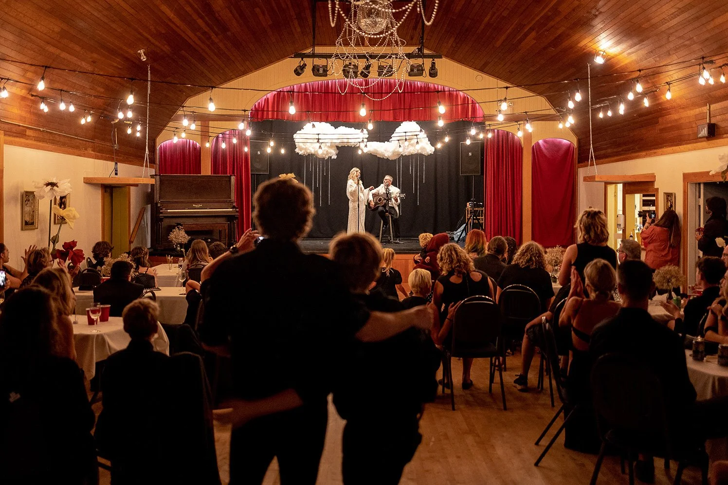 People sitting at tables watching a musical performance on stage with two singers, one playing guitar, decorated with clouds hanging from ceiling and string lights, in a wooden hall with red curtains.