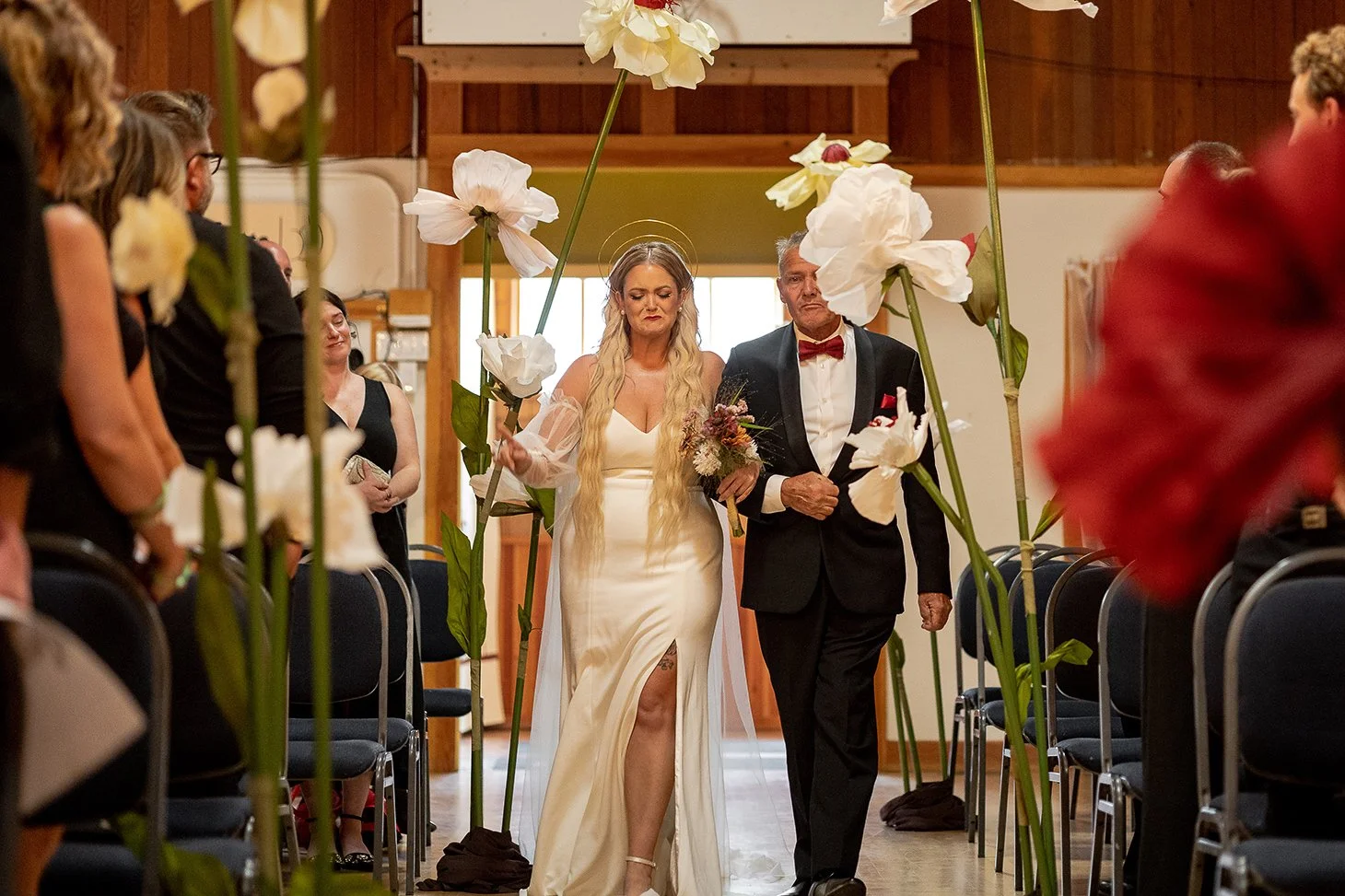A bride and an older man, possibly her father, walk down the aisle at a wedding ceremony, surrounded by large white flowers on tall green stems and seated guests.