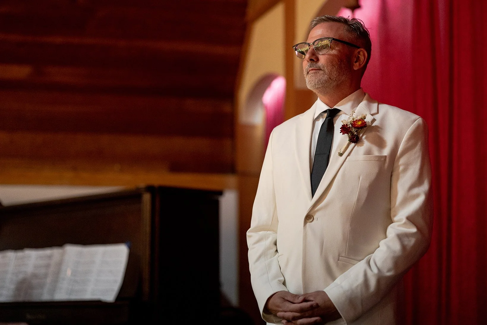 A man in a white tuxedo with glasses and a boutonniere stands with his hands clasped, looking serious, in front of a red curtain and a piano with sheet music.