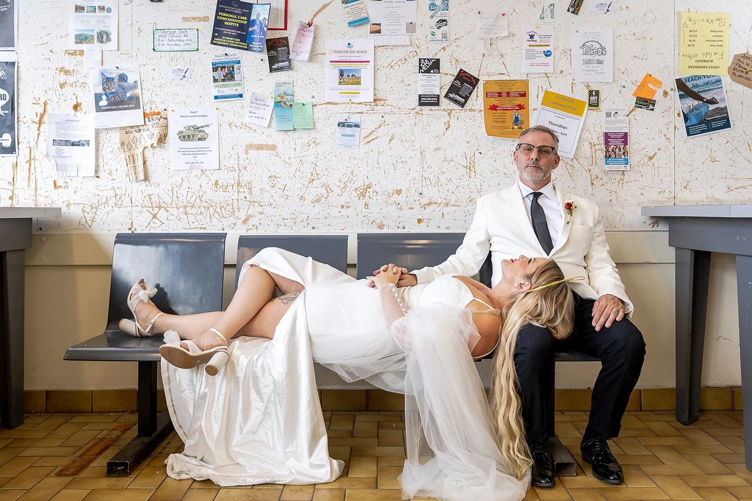 A bride in a wedding dress and heels reclines with her head on a man's lap, who is wearing a white suit and glasses, seated on a bench in a room with a busy, graffiti-marked wall and various flyers and papers pinned above.