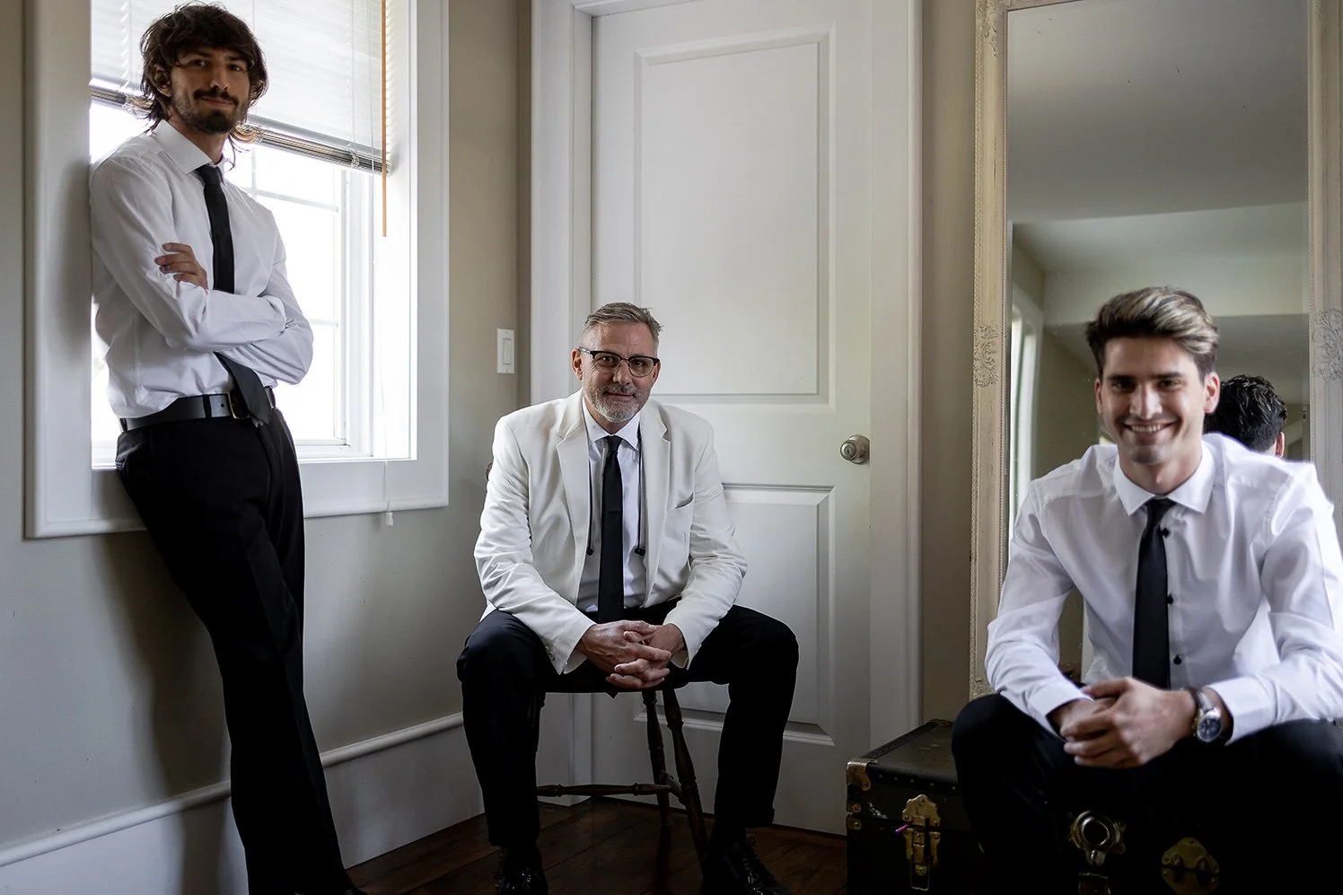 Three men dressed in white shirts and black ties, posing indoors near a mirror and a window. One man is standing with arms crossed, the second is seated on a stool, and the third is sitting on a suitcase, all smiling.